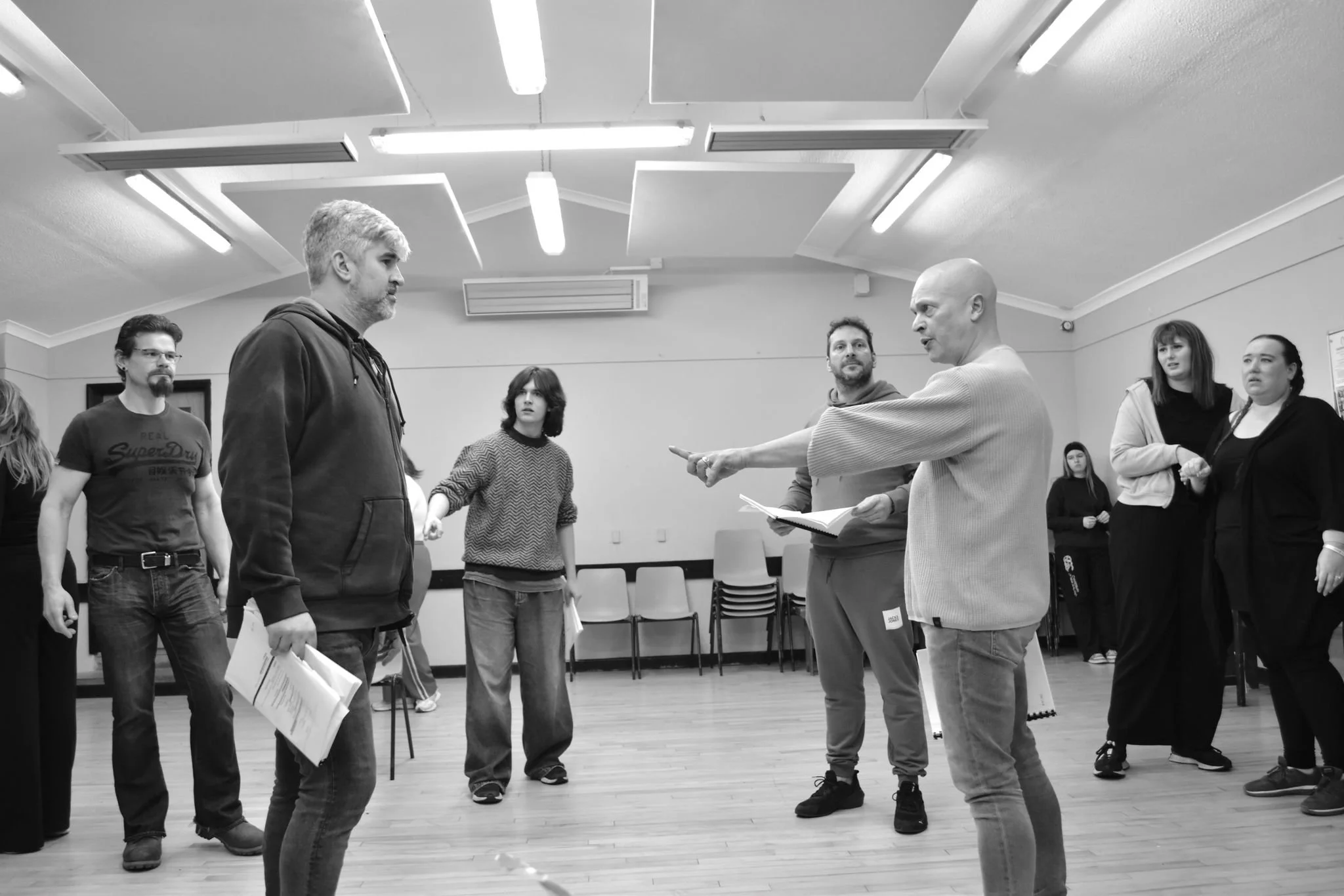 The image shows a group of people in a room during a rehearsal or acting workshop, with an instructor pointing and giving directions to a participant in front.