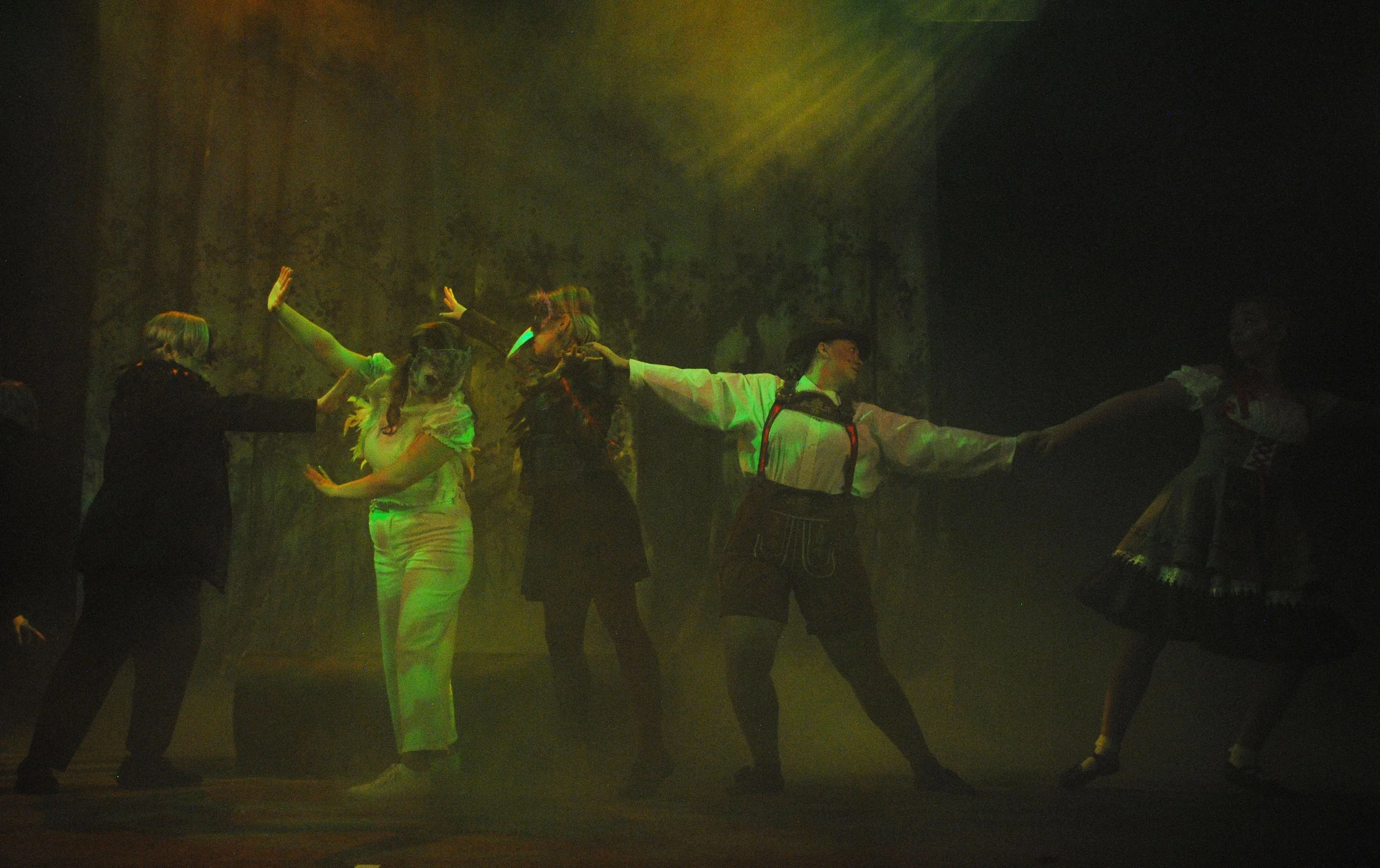 Stage performance with five women, some in traditional and some in modern costumes, engaged in a choreographed dance or theatrical act under green and yellow lighting with a dark background.