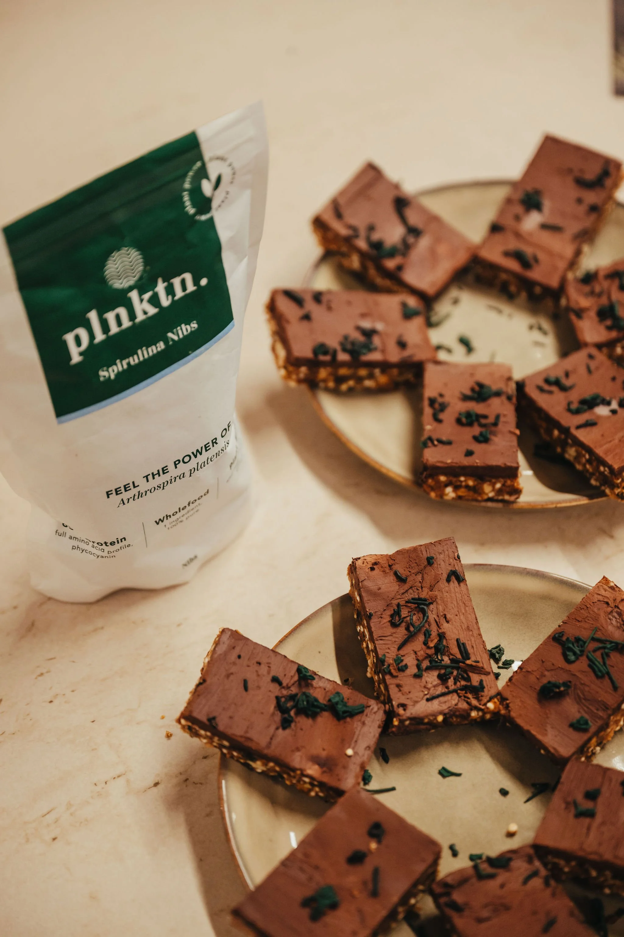 Close-up of chocolate squares topped with green herbs and sprinkles on ceramic plates, with a bag labeled 'plnktn spirulina nibs' nearby.