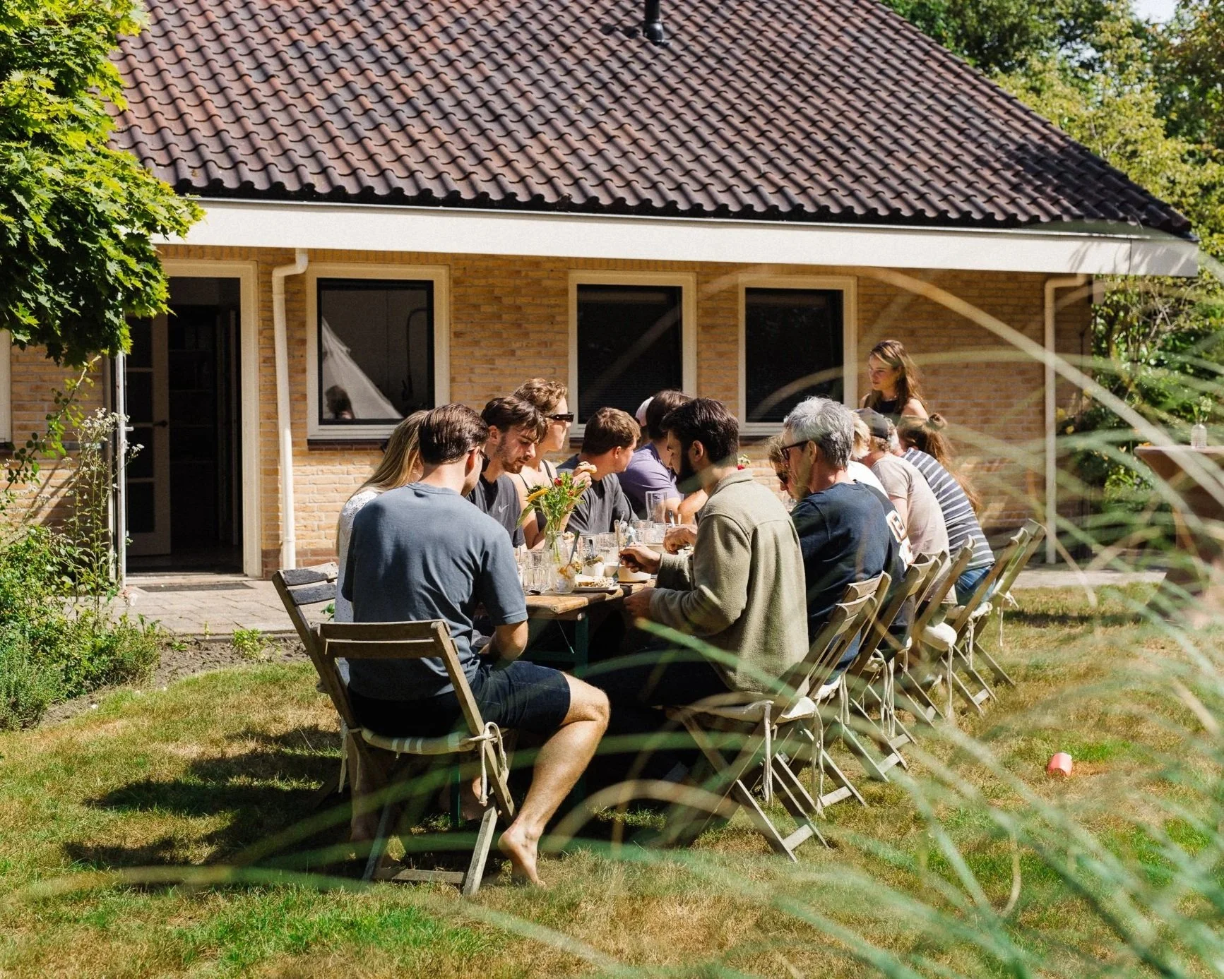 Een groep mensen zit in een tuin aan een lange tafel op een zonnige dag en eet samen ontbijt