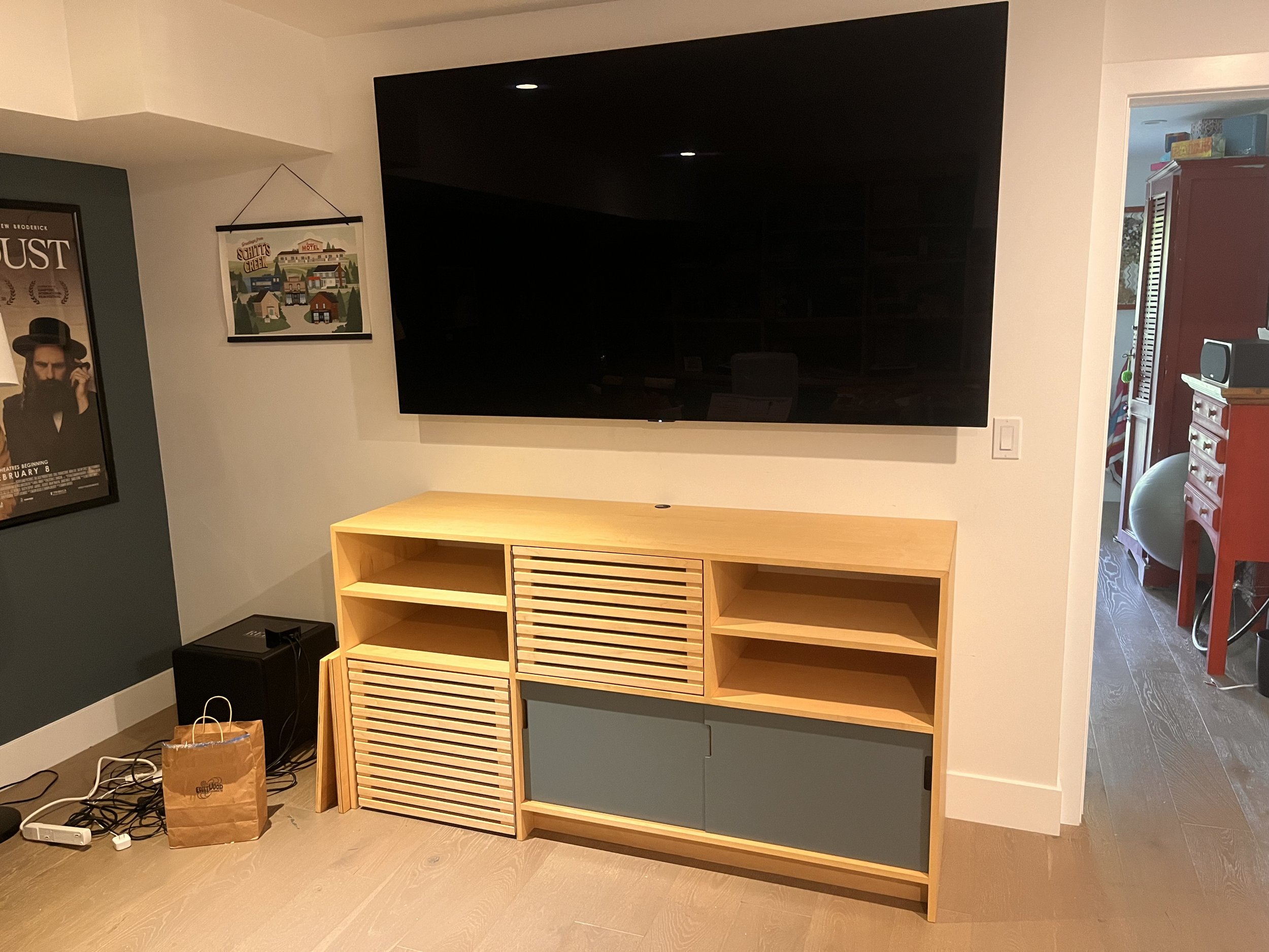 A wooden storage area below a wall mounted tv in a residential house. It has thin shelves and pull out draws.