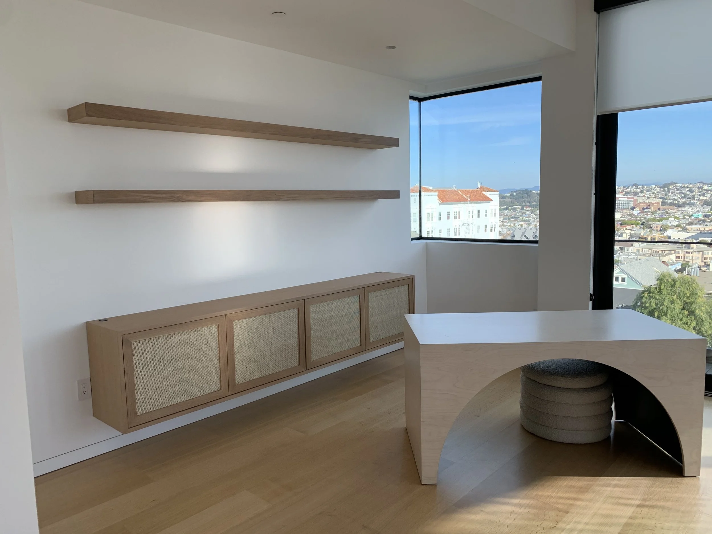 Wall shelves and cupboard with fabric doors are mounted on a wall in a residential house. The table in the middle of the room is a bridge like design.