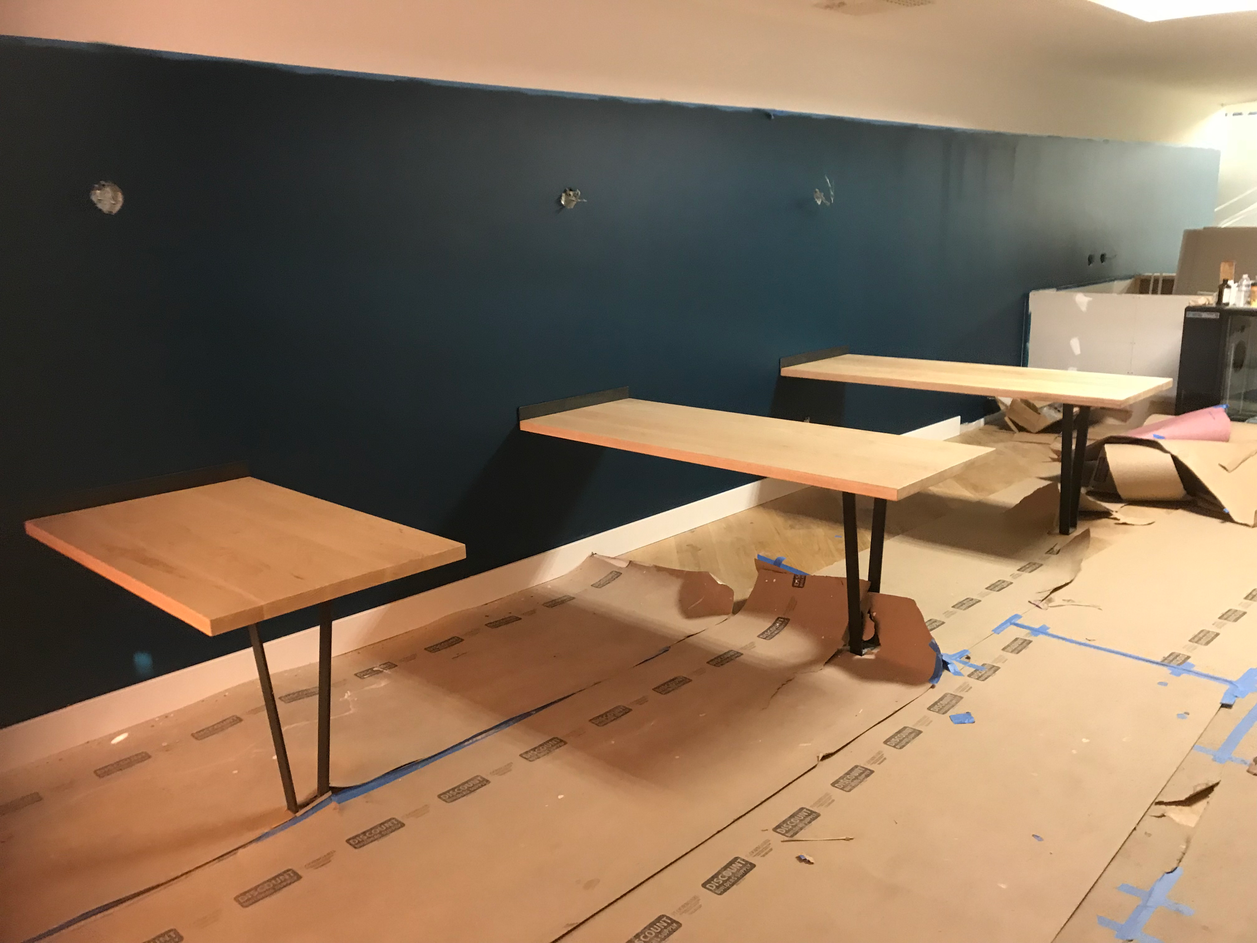 Custom wooden tables with metal legs designed for booths being installed in an empty room.