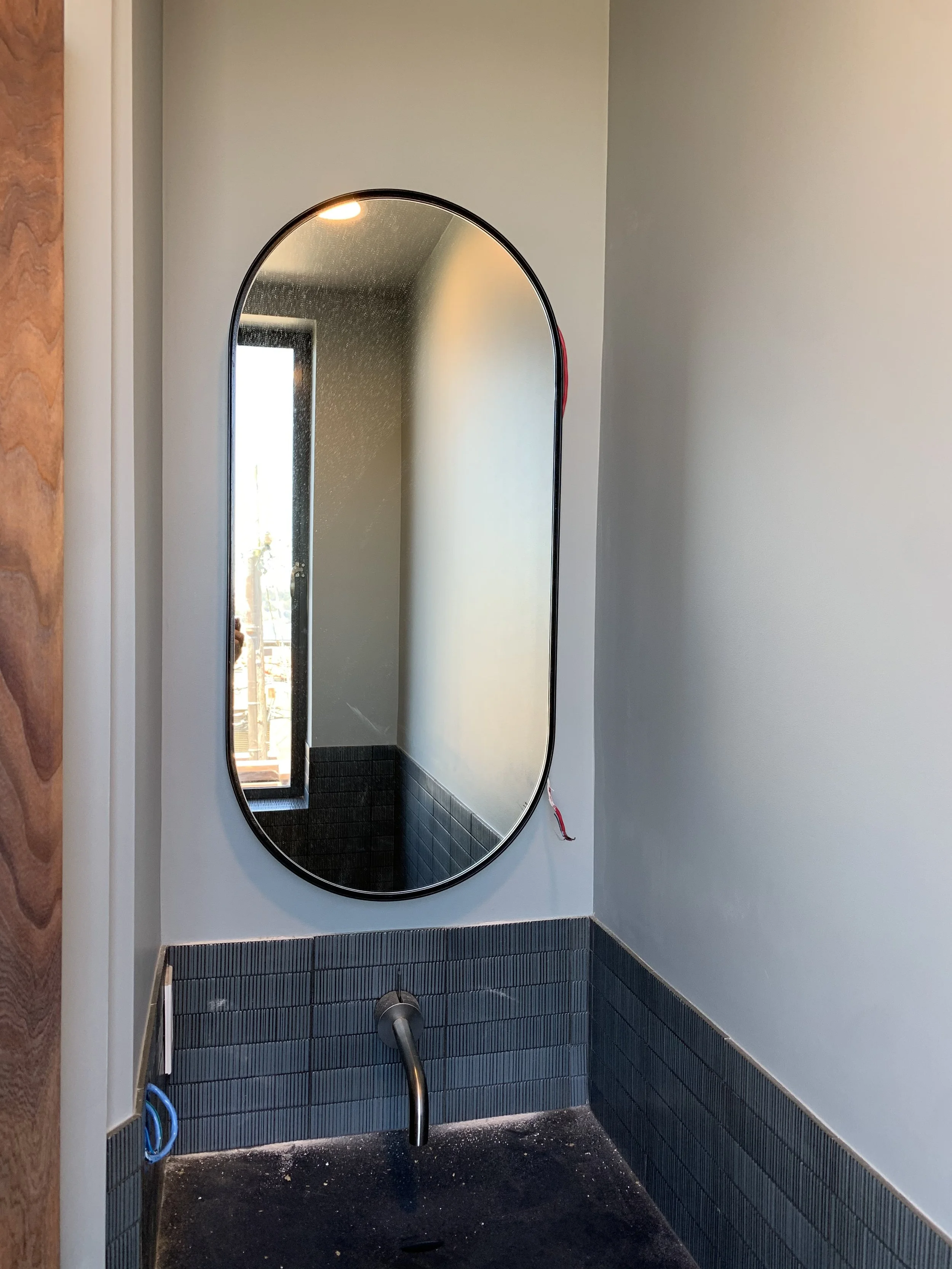 A tall pill shaped mirror in a residential bathroom designed to fit in the narrow area the sink is in.