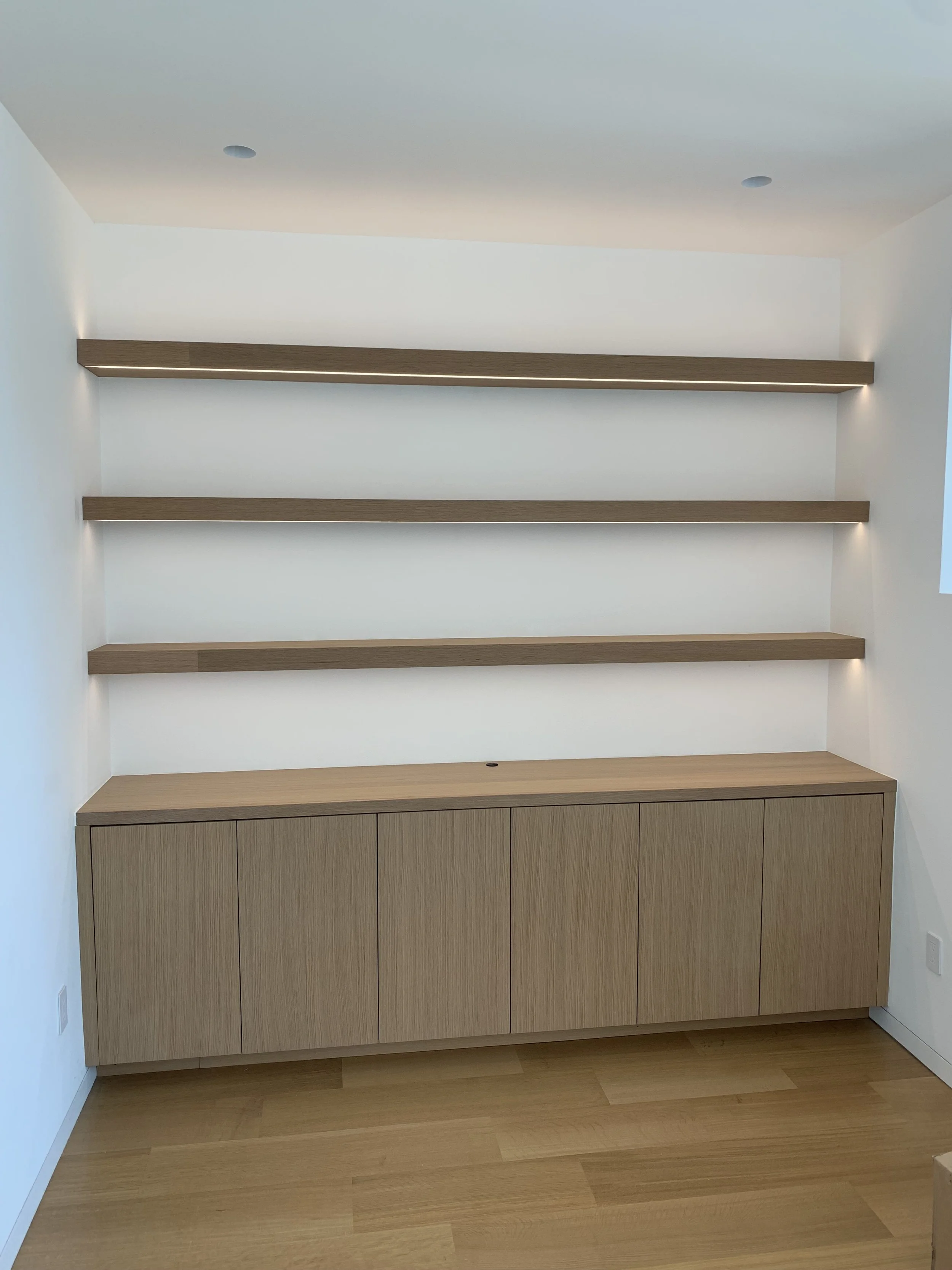 Wooden drawers spanning the length of the wall with 3 wooden shelves above the drawers. The shelves have lights above and below them.