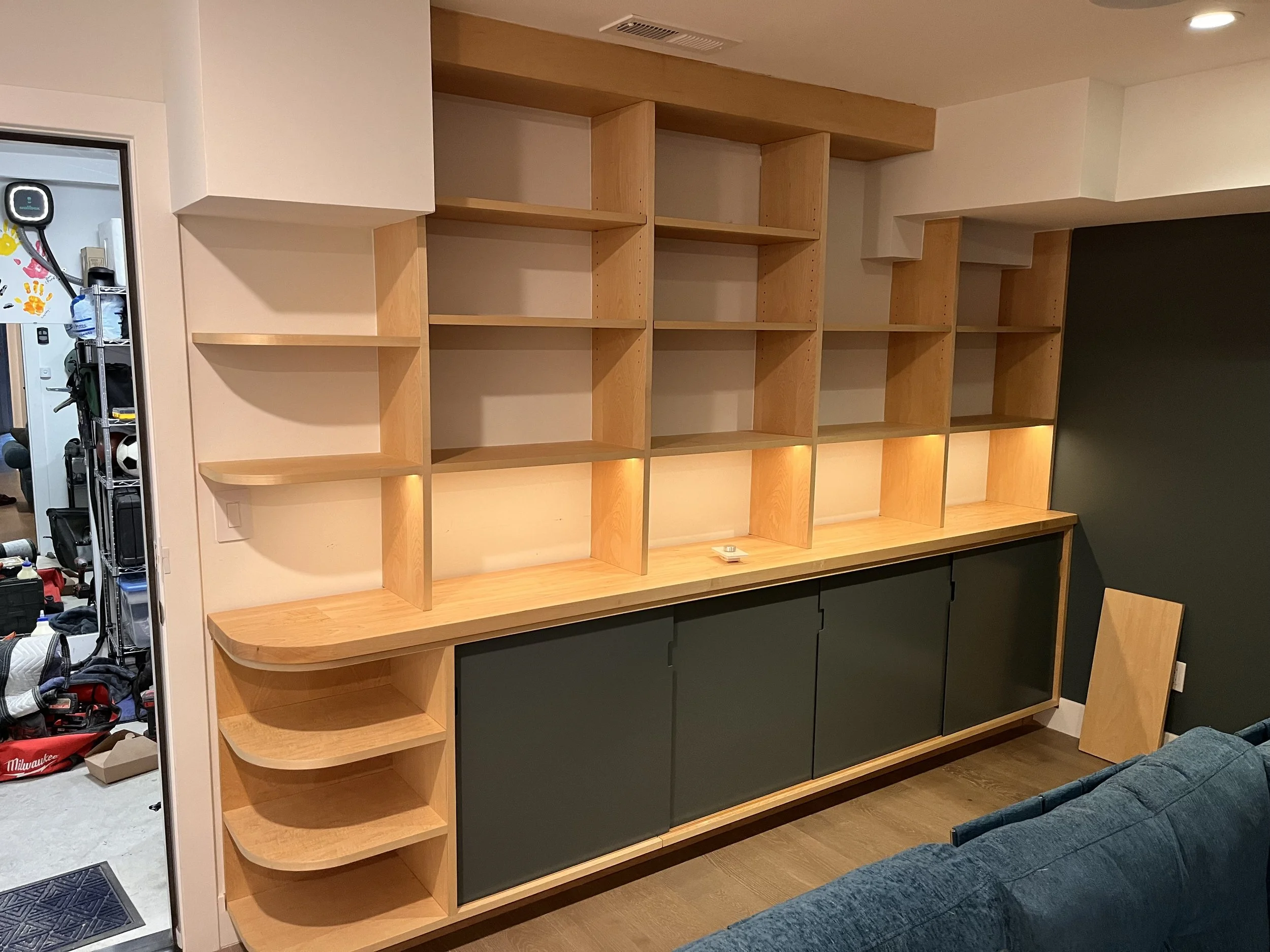 A custom wooden storage/shelving space along the wall, shelves at the left side are rounded off. The shelves along the top of the wall are designed to fit in-between the odd roof shape.