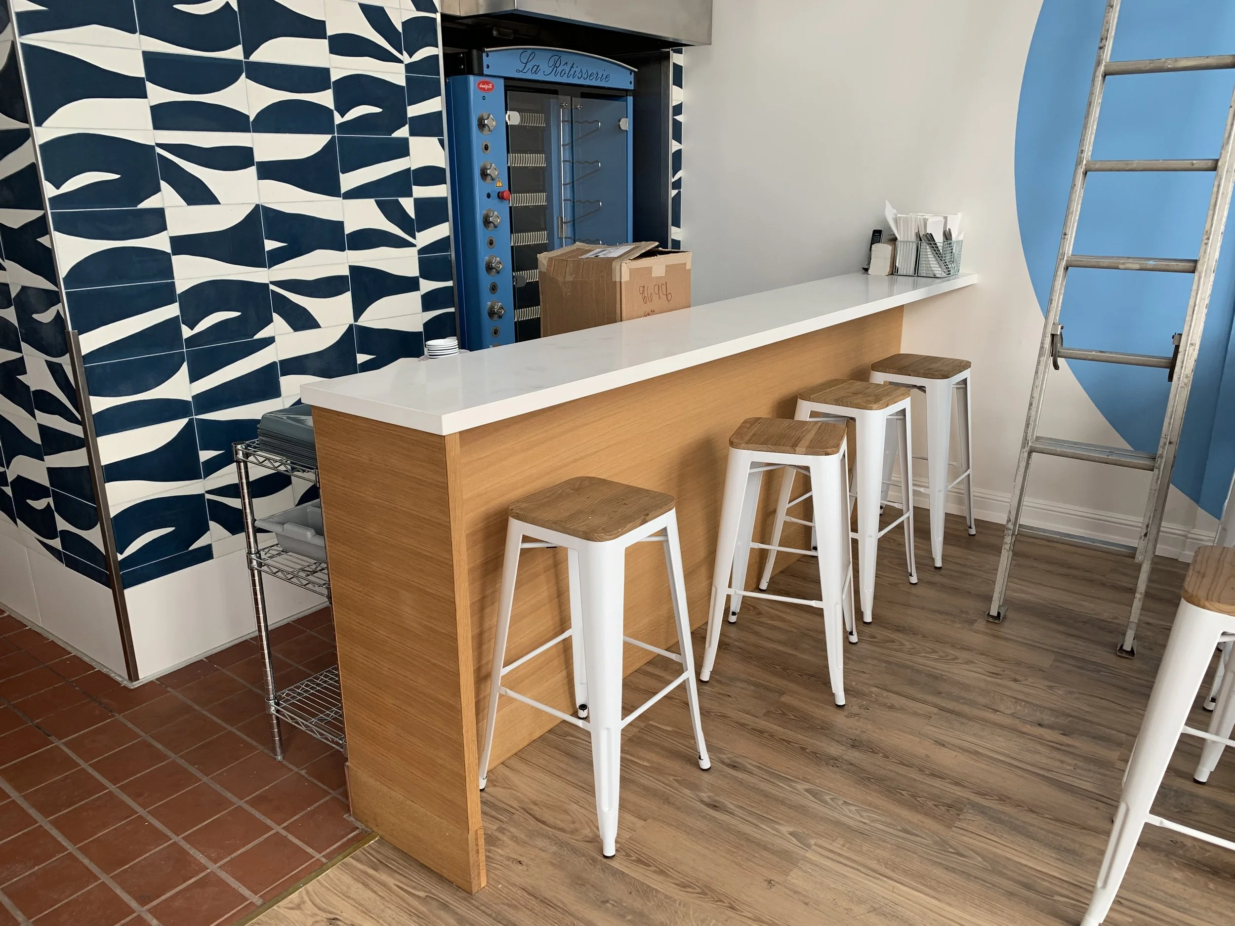 A counter with a wooden base and a white plastic counter top. The counter is designed with a seating area for custom made stools, they have a wooden seat with white, metal legs.