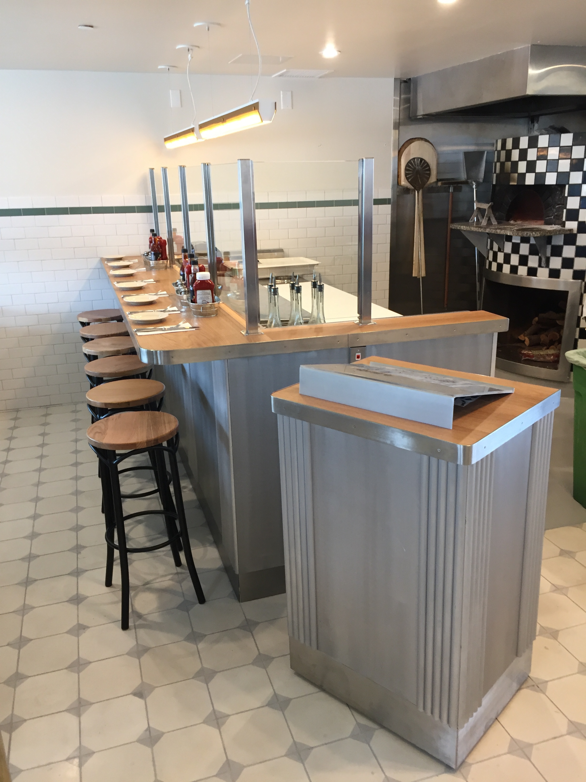 A large bar table with an area for stools. The bar table has a wooden work top with a metal covering over the bottom. Next to the table is a host stand made of and in the same style of the other table.