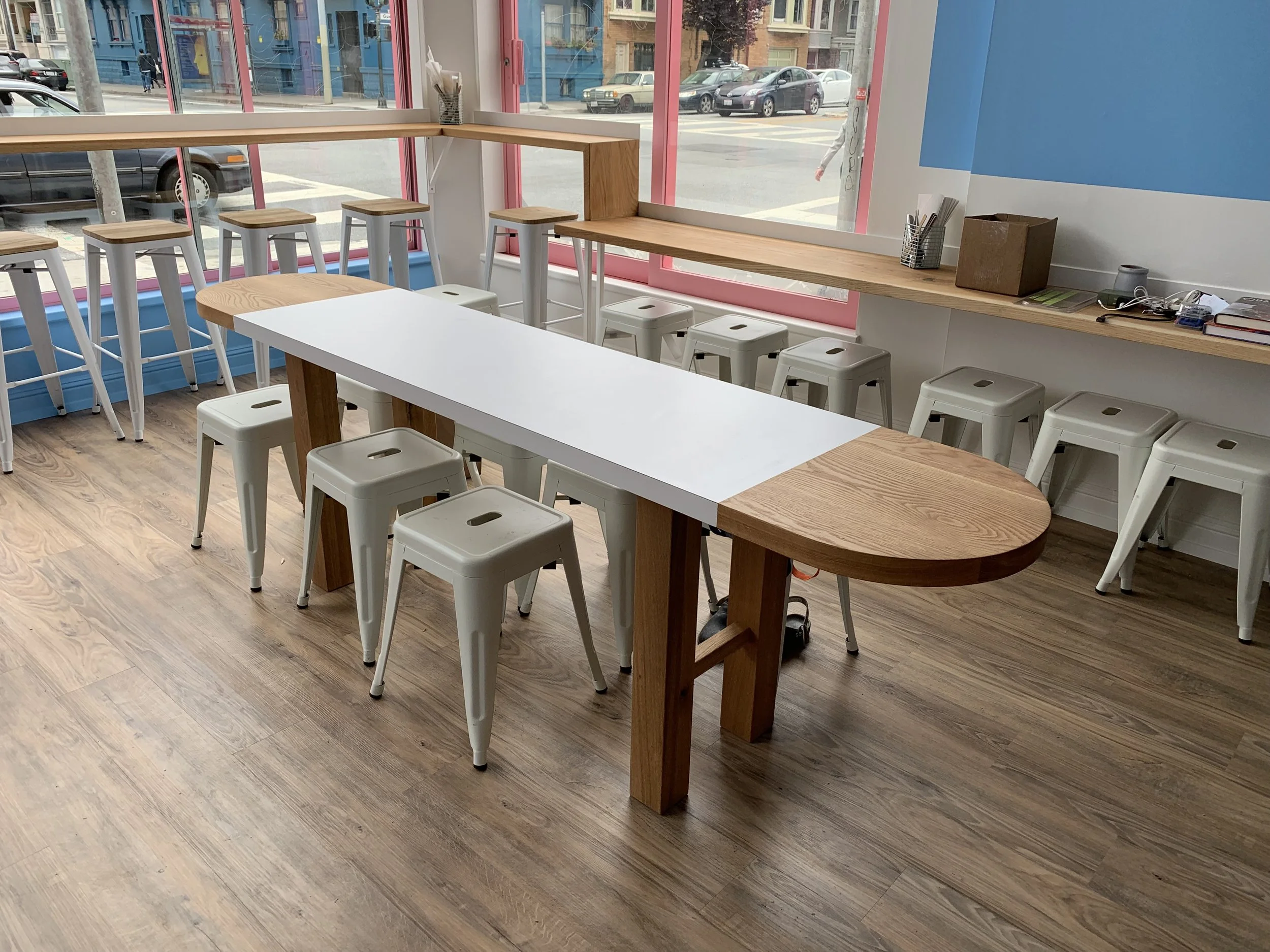 A long pill shaped table in the middle of the room with a plastic section in the middle for eating. Along the edge of the room is a continuous wooden counter for eating either space for custom made stools below.