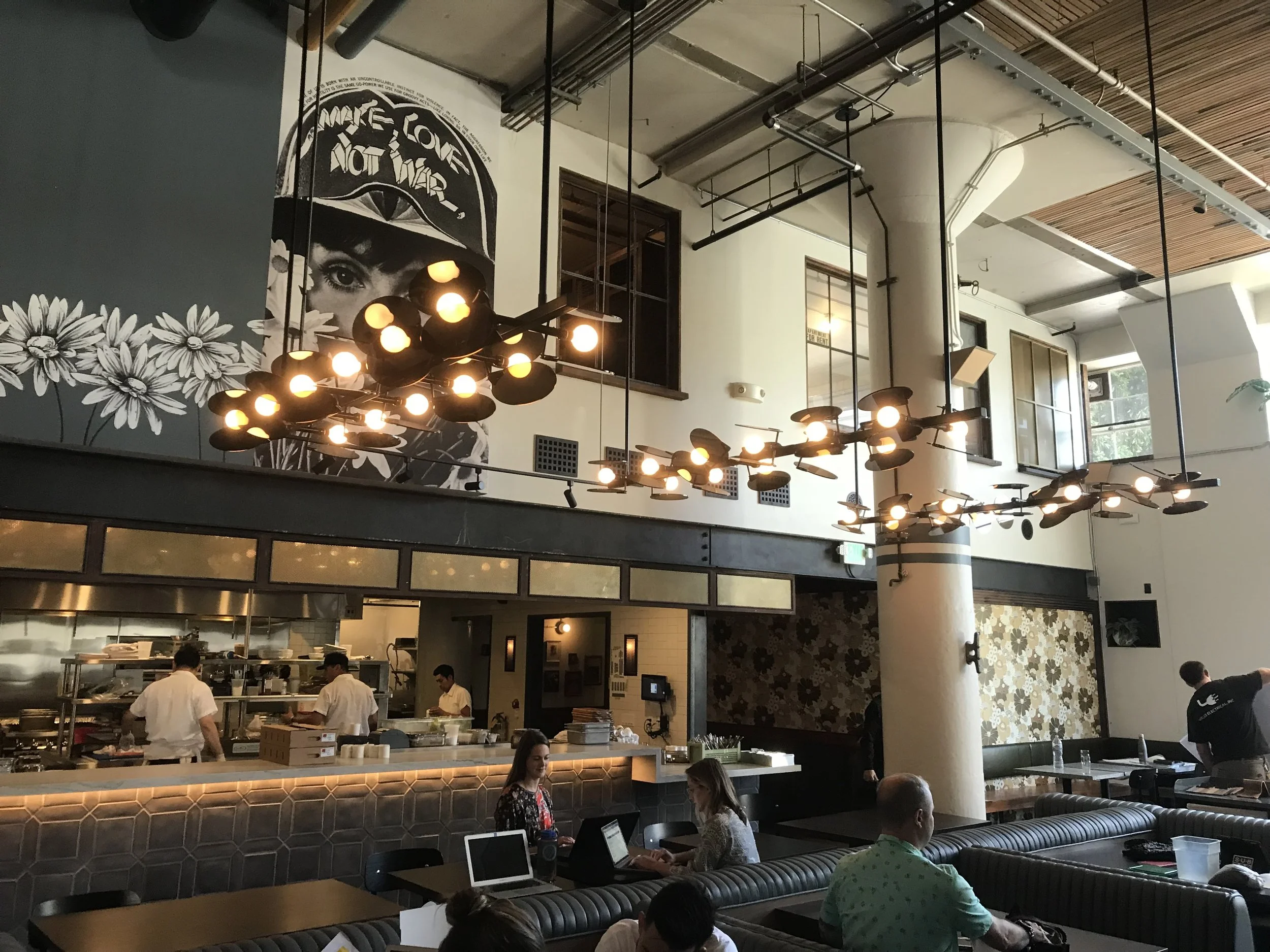A wide shot from the seating area of a restaurant looking towards the open kitchen area. Lights in the shape of hour glasses can be seen hanging fro the roof. The seating area has custom seating and wooden table tops in the centre of the restaurant.