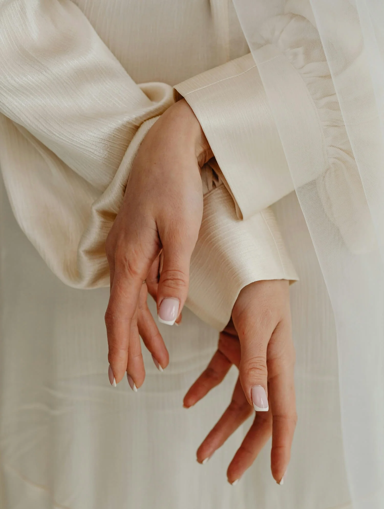 Close-up of a person's hands with manicured nails, crossing in front of their torso, dressed in cream-colored, silk-like fabrics.