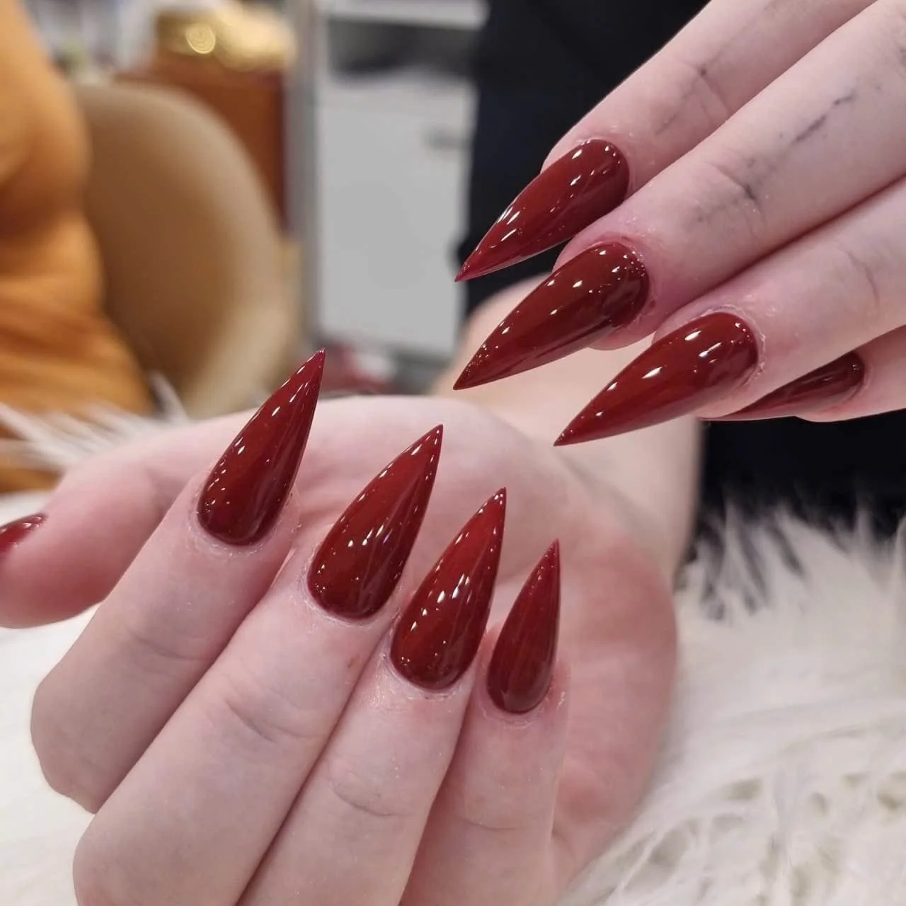 Close-up of a hand with gold manicured nails, wearing rings, resting on clothing with black and white fabric, with a necklace visible in the background.