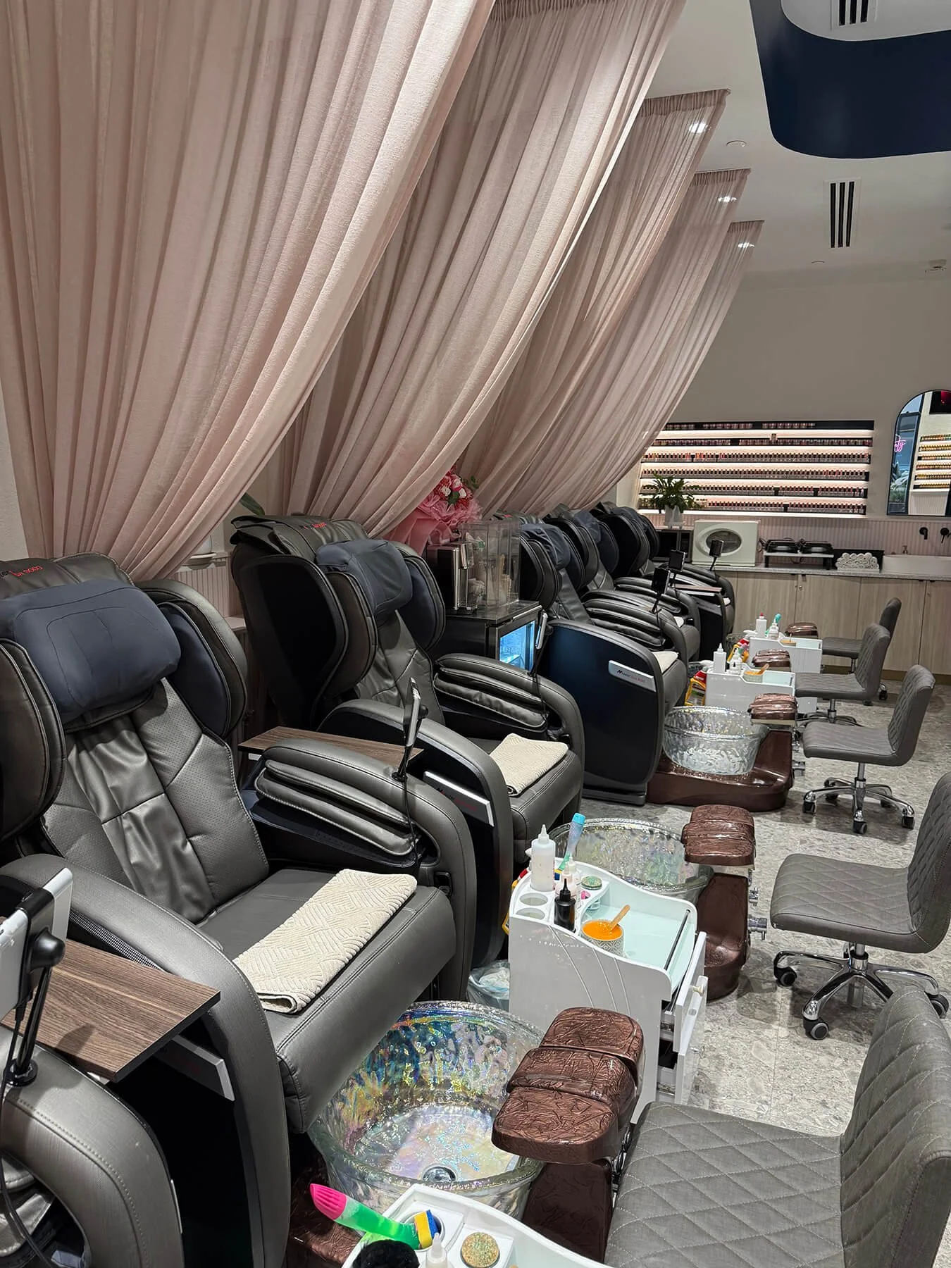Row of massage chairs at a nail salon with manicure stations, nail polish displays, and curtains in the background.