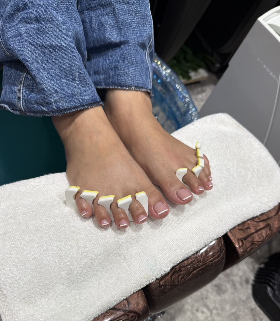 Close-up of a person's bare feet on grass, surrounded by white daisies, with hands gently holding the feet and daisies on the toes, wearing a brownish-orange garment.