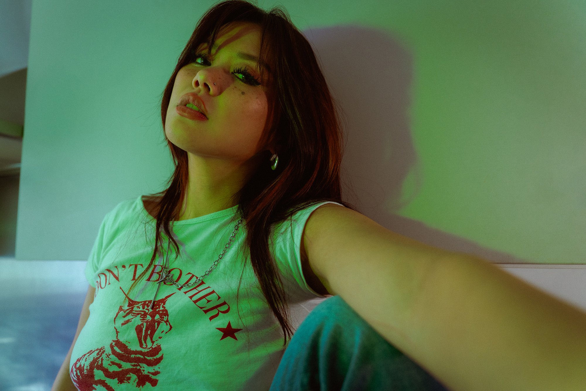A young woman with shoulder-length dark hair, fair skin, and freckles, wearing a white t-shirt with red graphic and text, jewelry, and makeup, taking a selfie against a plain background illuminated with green lighting.
