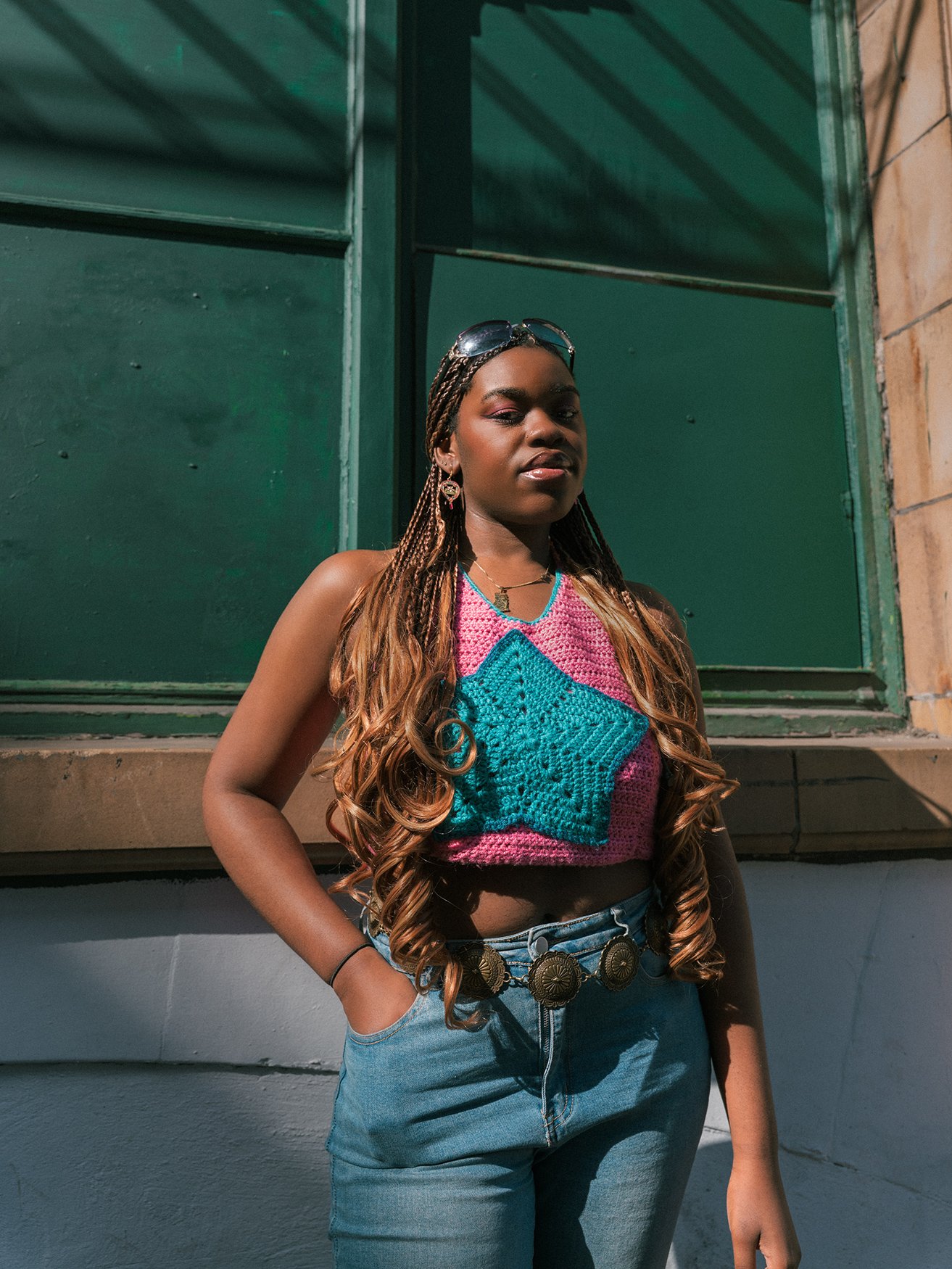 A woman with long, wavy hair, wearing sunglasses on her head, a colorful crocheted crop top with a star design, light blue jeans with a decorative belt, standing outdoors in front of a green building with large windows.