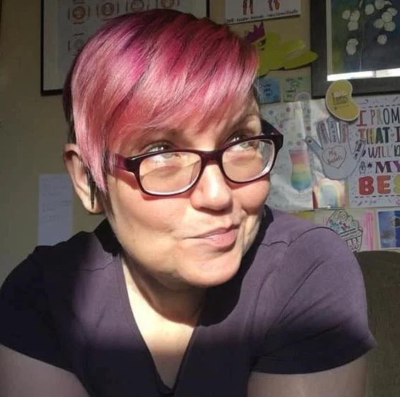 A woman with glasses and pink hair looks away from the camera with a slight smile on her face