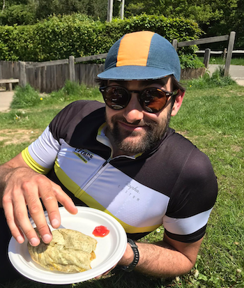A man with a beard and cycling jersey on enjoys a pasty while relaxing on some grass