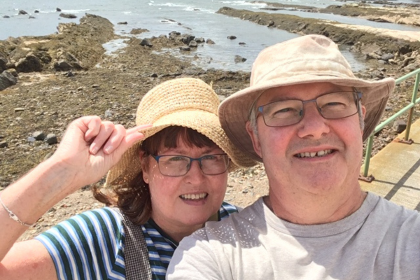 A man and woman in sun-hats smiling on a walk, behind them is the sea and some rocks
