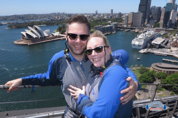 A man and a woman in grey and blue jackets posing on top of sydney harbour bridge
