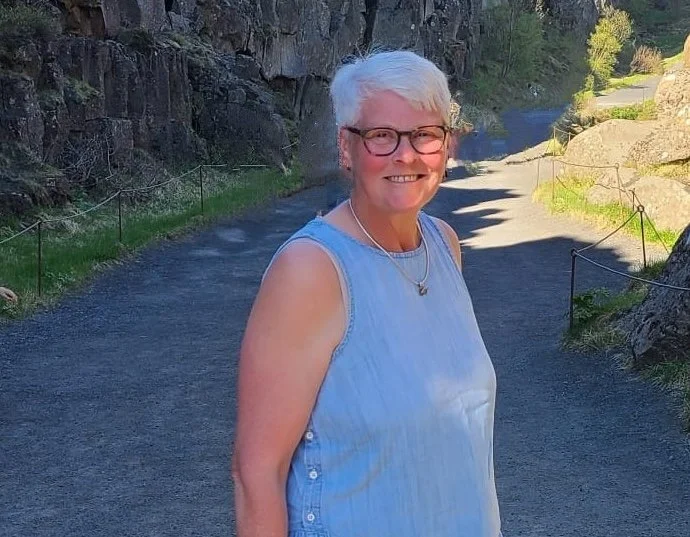 A photo of a woman in her 60s with a blue sleeveless top on, smiling during a walking in nature