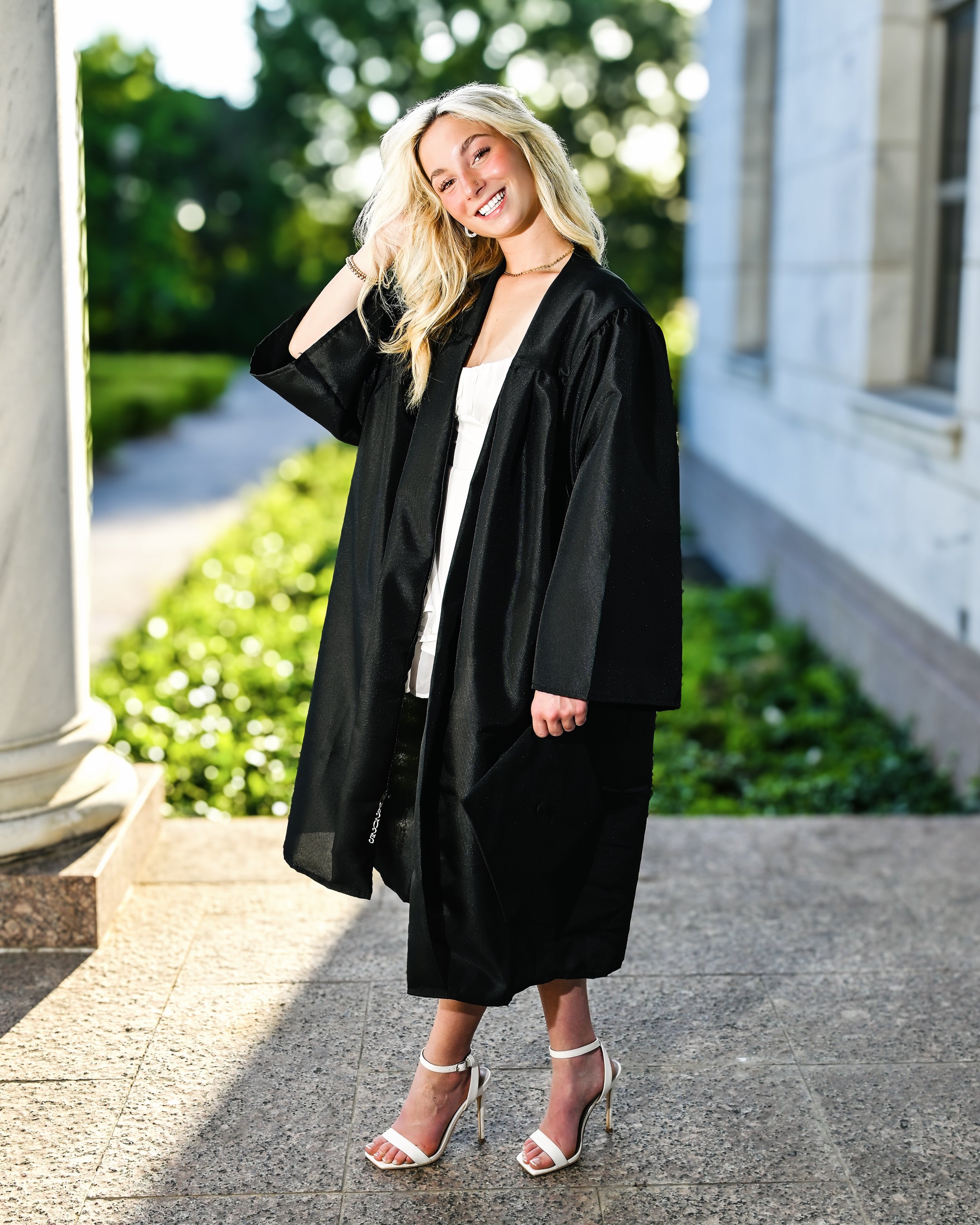 A young woman in a graduation gown smiling outside on a sunny day, holding a black cap.