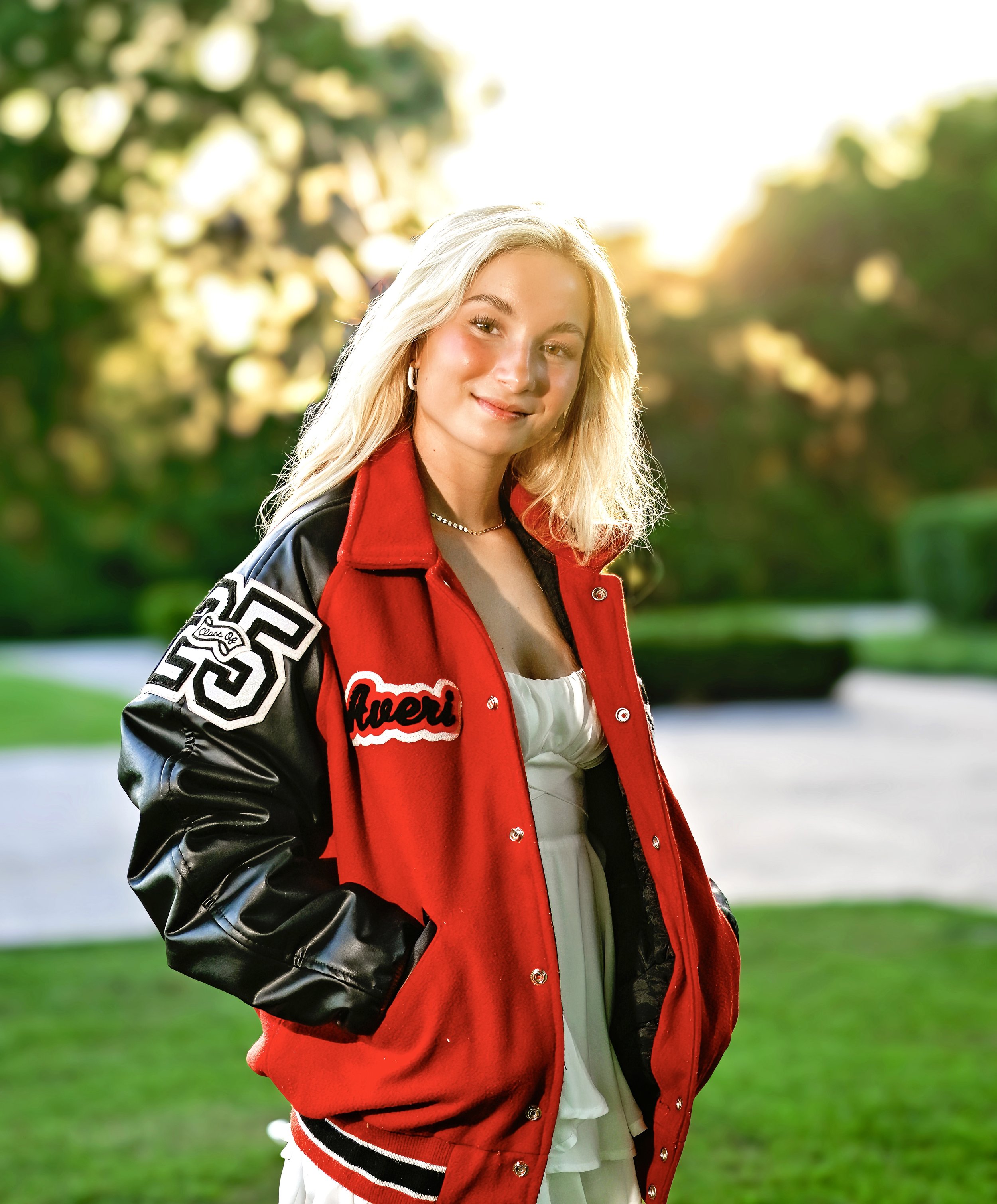 A young woman with blonde hair, wearing a red and black letterman jacket, stands outdoors on a sunny day with a blurred green park background.