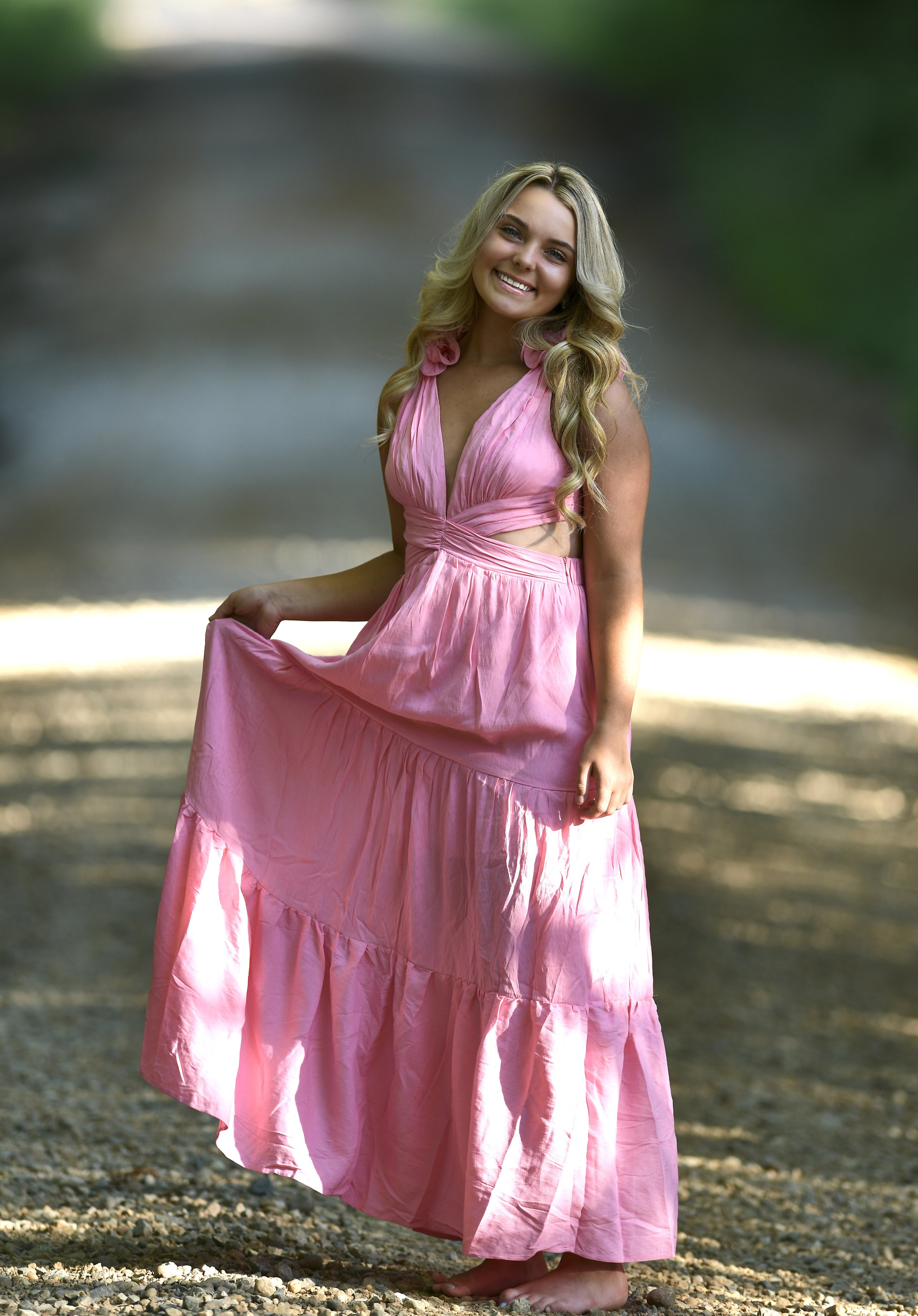 A young woman with long blonde hair wearing a pink maxi dress, standing barefoot on a gravel path in a wooded area, smiling at the camera.