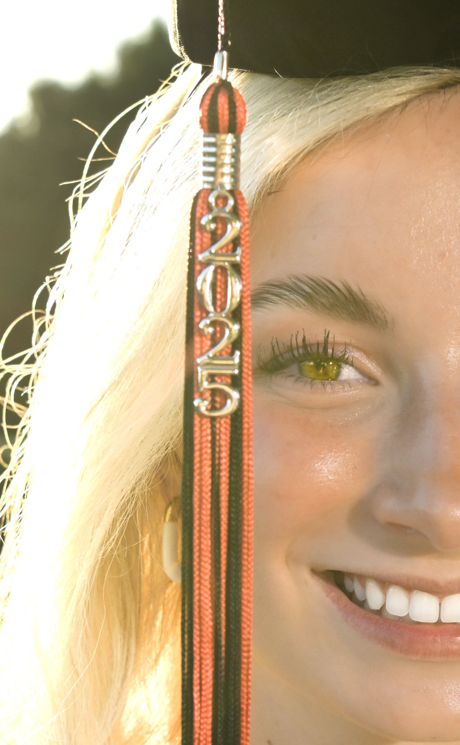 Close-up of a young woman's face smiling with green eyes, wearing a black graduation cap with a red and black tassel that has the year 2023 on it, partially covering one eye, with a natural outdoor background.