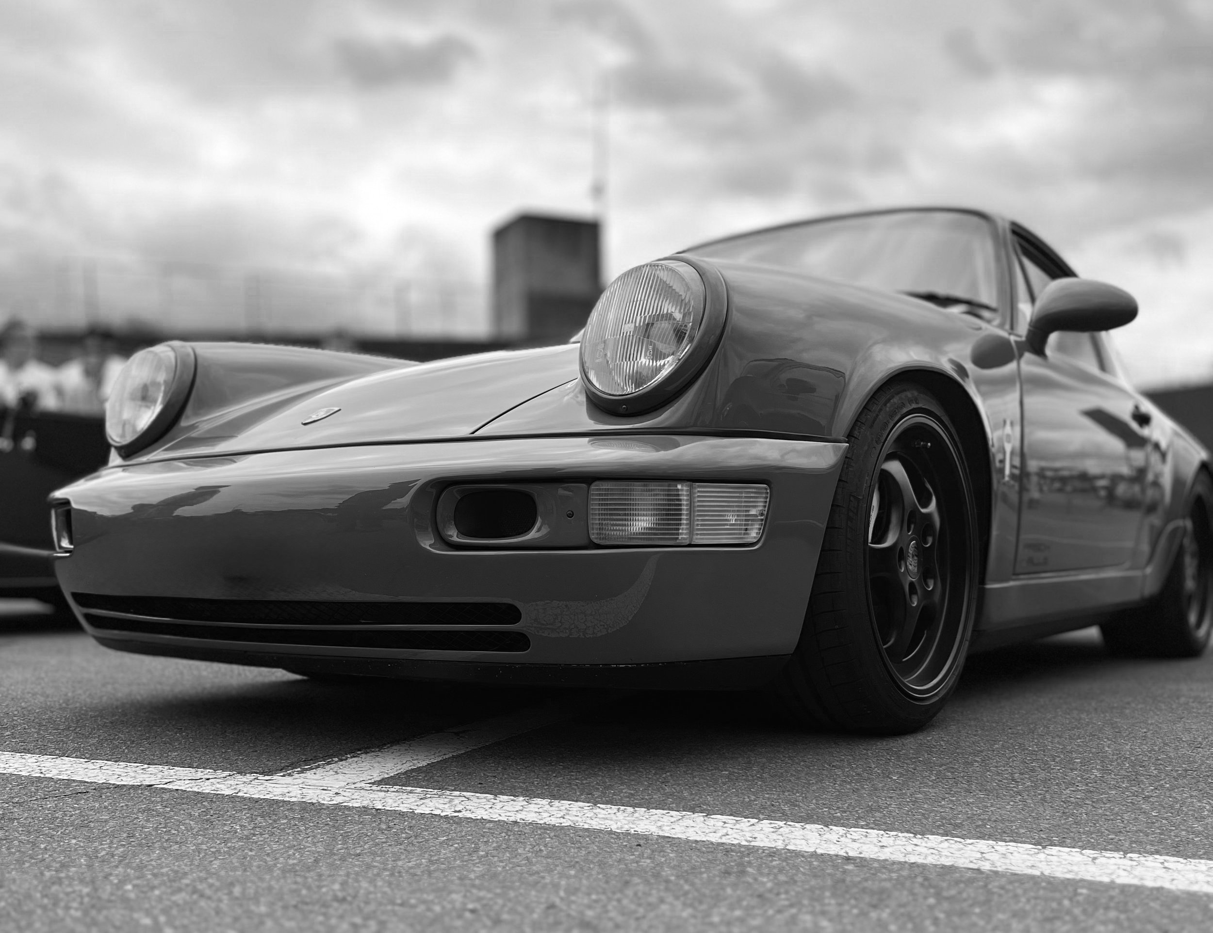Black and white photo of a classic Porsche sports car parked in a lot with a cloudy sky above.
