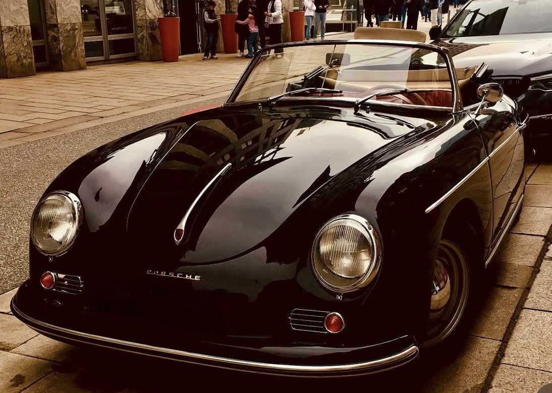 A vintage black Porsche convertible parked on a city street with people and modern cars in the background.