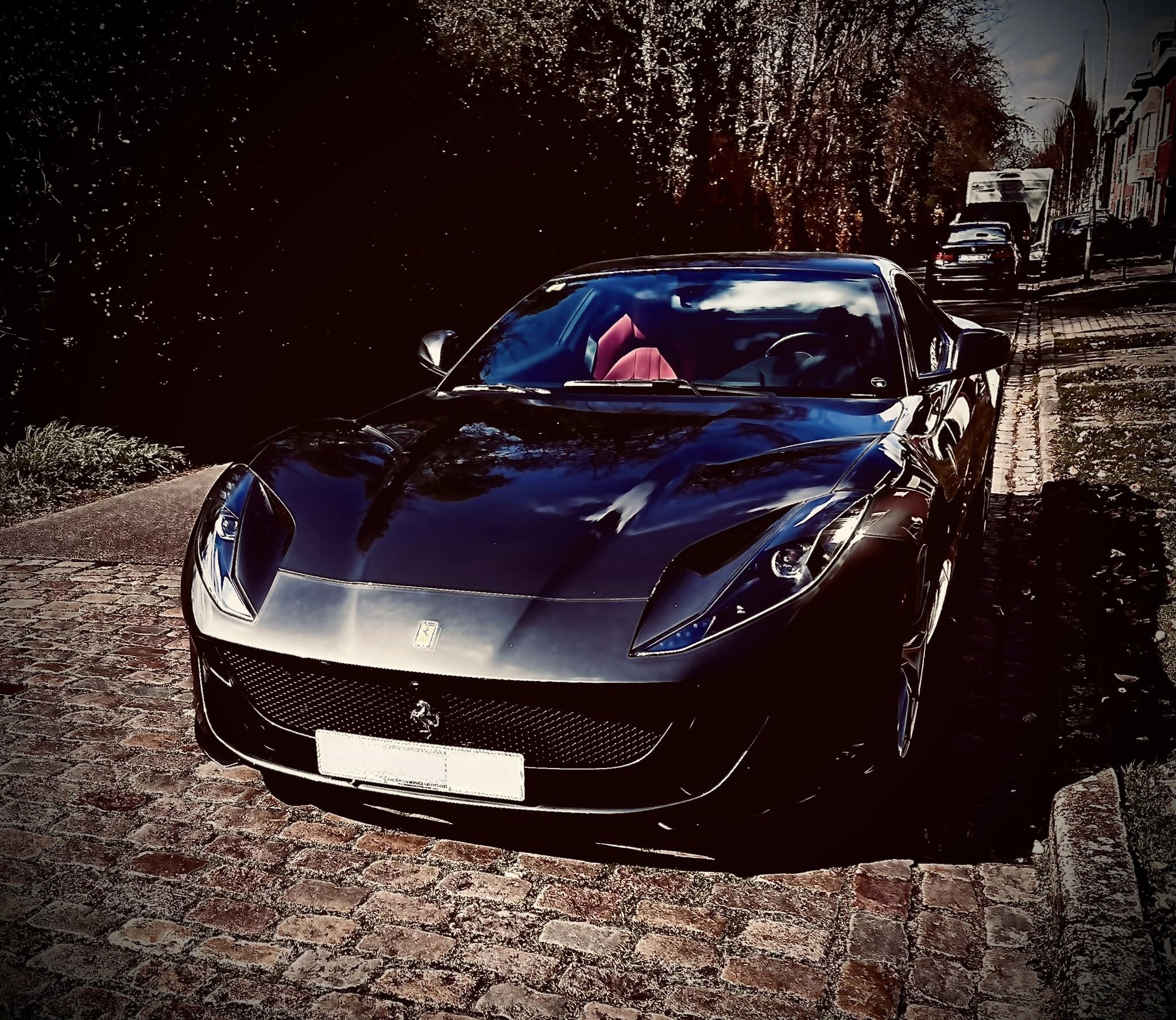 Black Ferrari sports car parked on a cobblestone street with trees and other cars in the background.