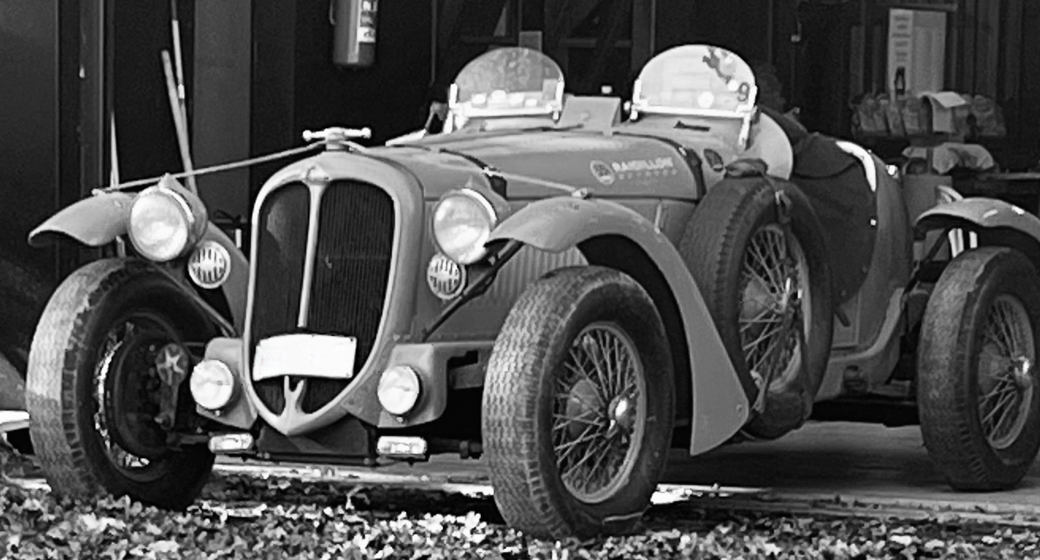 A vintage race car with wire wheels, round headlights, and an open cockpit, parked outdoors in front of a garage, in black and white.