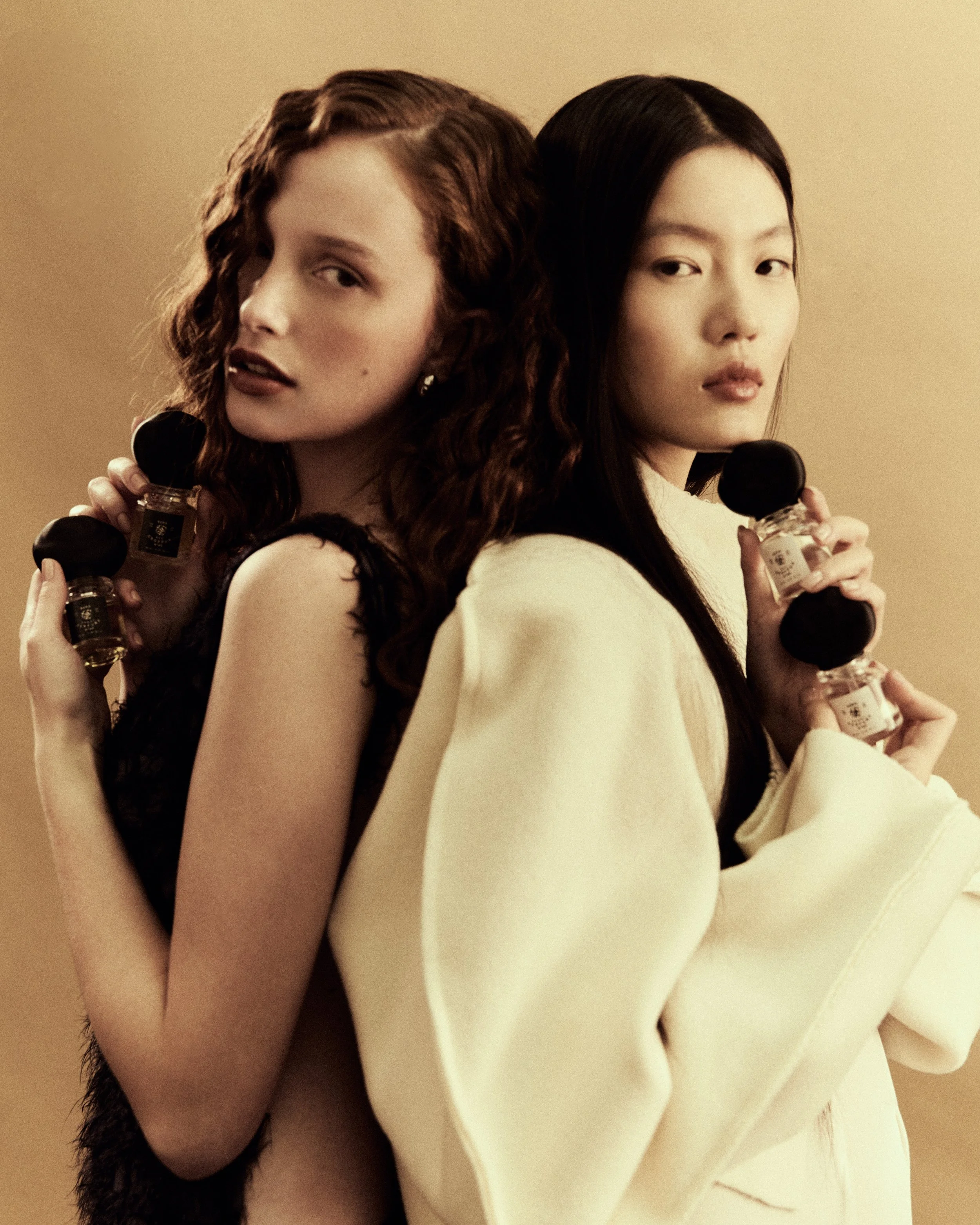 Due donne eleganti siedono indossando abiti e tengono bottiglie di profumo, con sfondo beige.