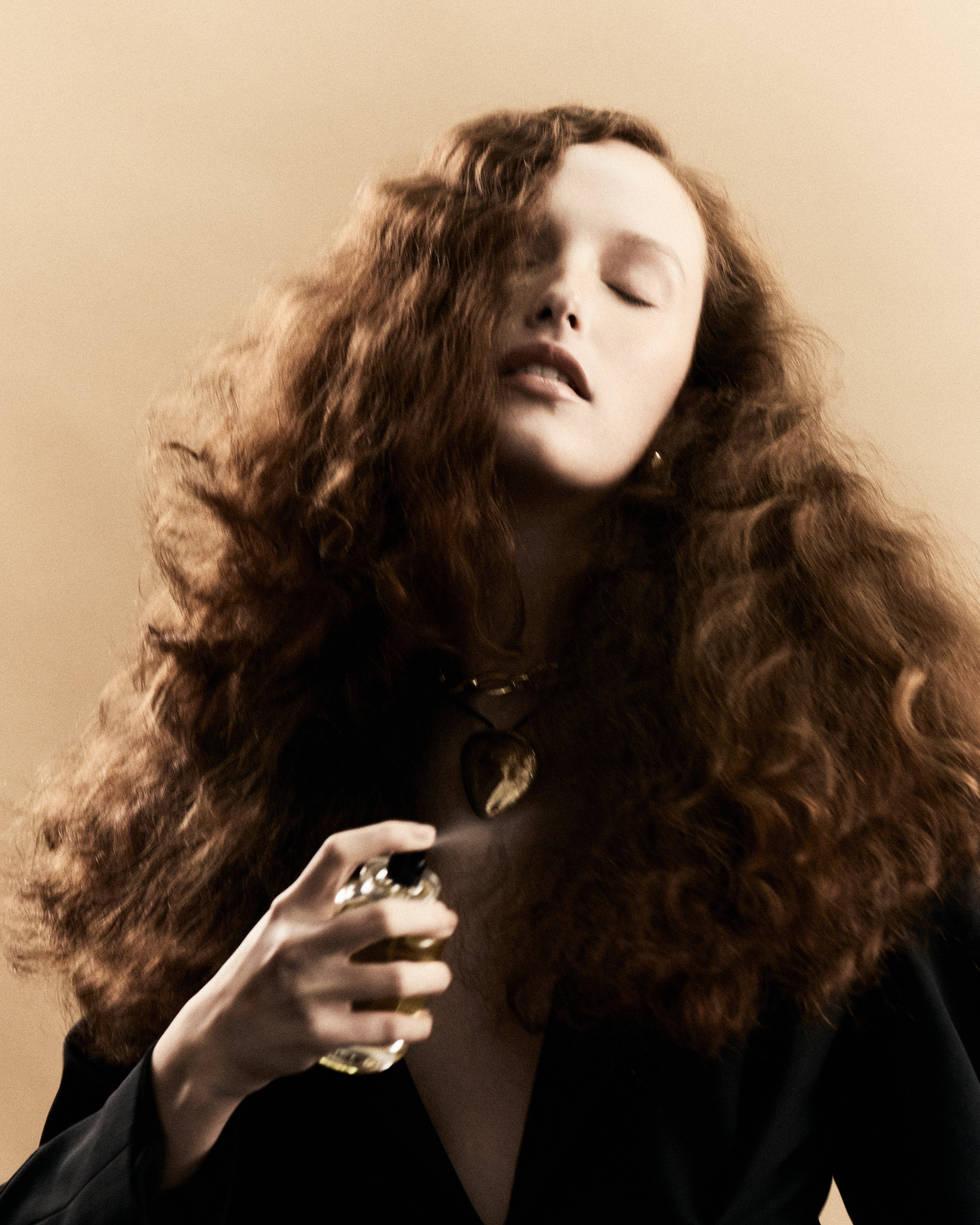 Una donna con lunghi capelli ricci e castani che tiene una bottiglia di profumo, indossa una collana con una grande cassa e un abito nero, sfondo neutro.