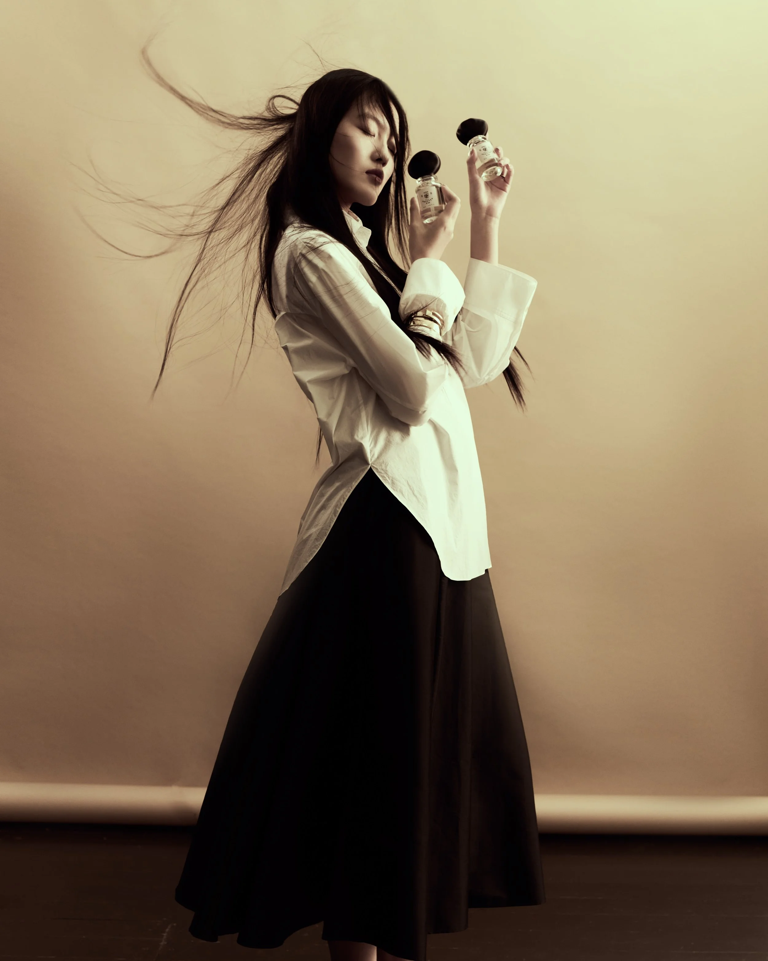 Donna con capelli lunghi e scuri tiene due bottiglie di olio o profumo, indossa una camicia bianca e una gonna lunga nera, sfondo neutro.