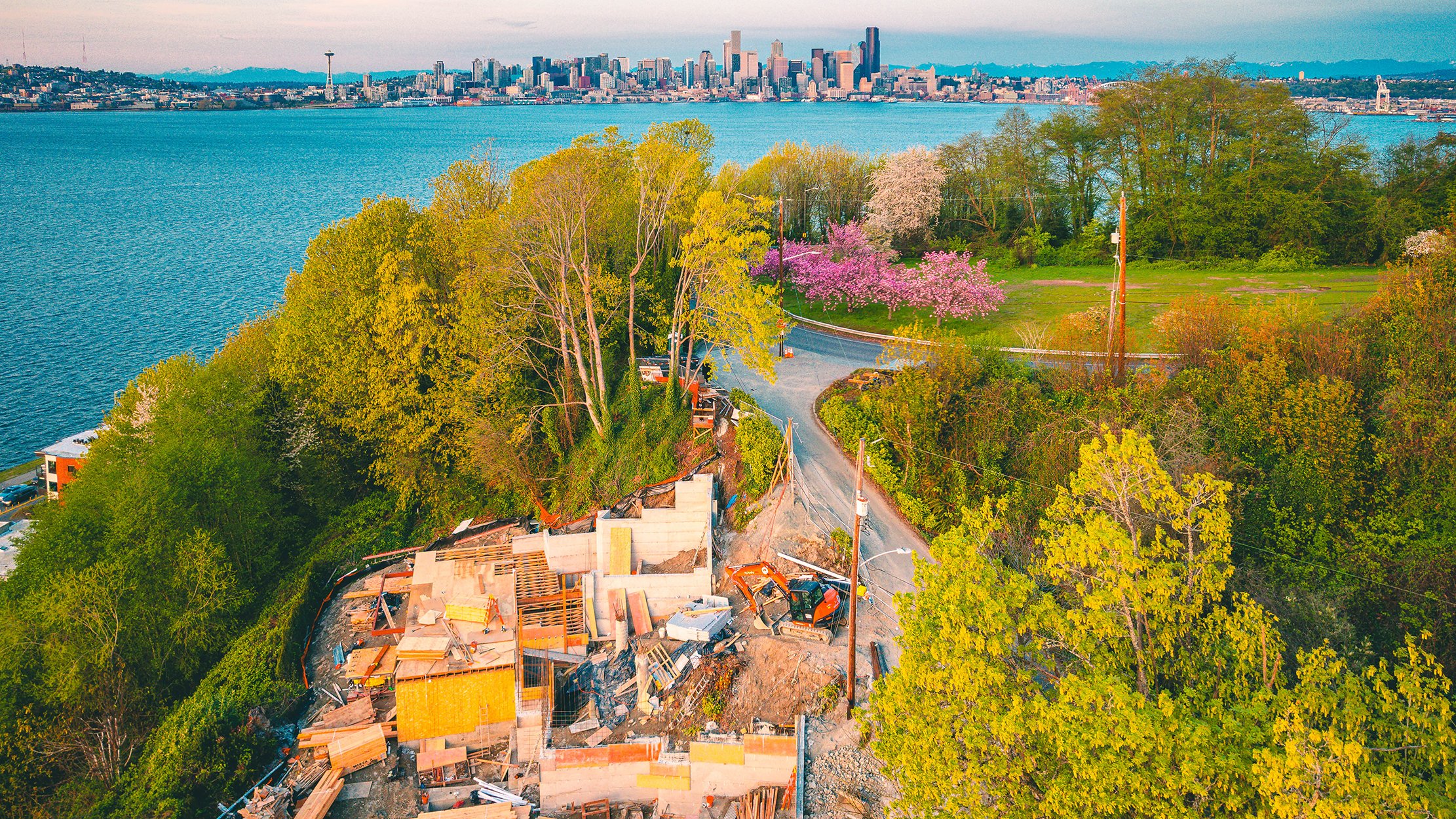 Aerial view of a construction site on a hillside with trees, overlooking a body of water and a city skyline in the background, during daytime.