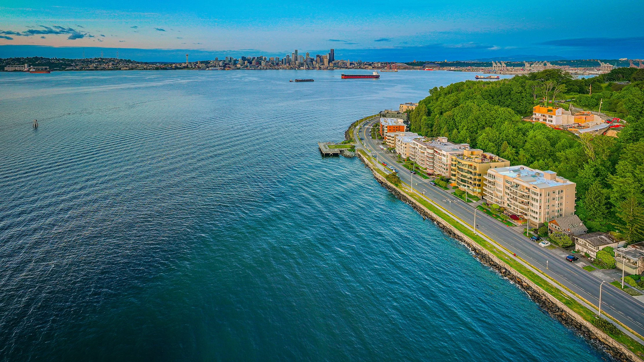 Aerial view of the Puget Sound with a road along the shoreline, apartment buildings, and lush green hills. The city skyline with tall buildings is visible across the water, with ships in the harbor and a cloudy sky overhead.