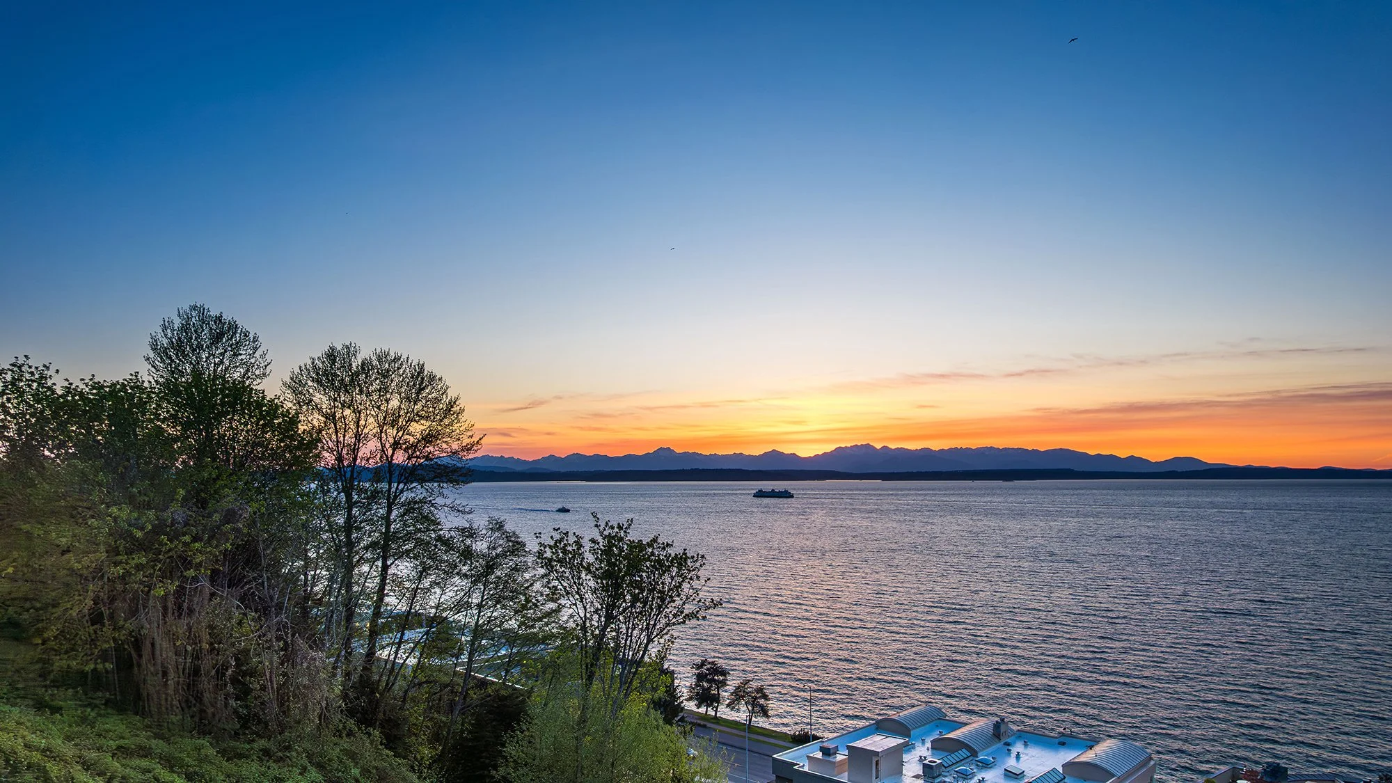Sunset over a body of water with mountain range in the distance, trees and a building in the foreground.