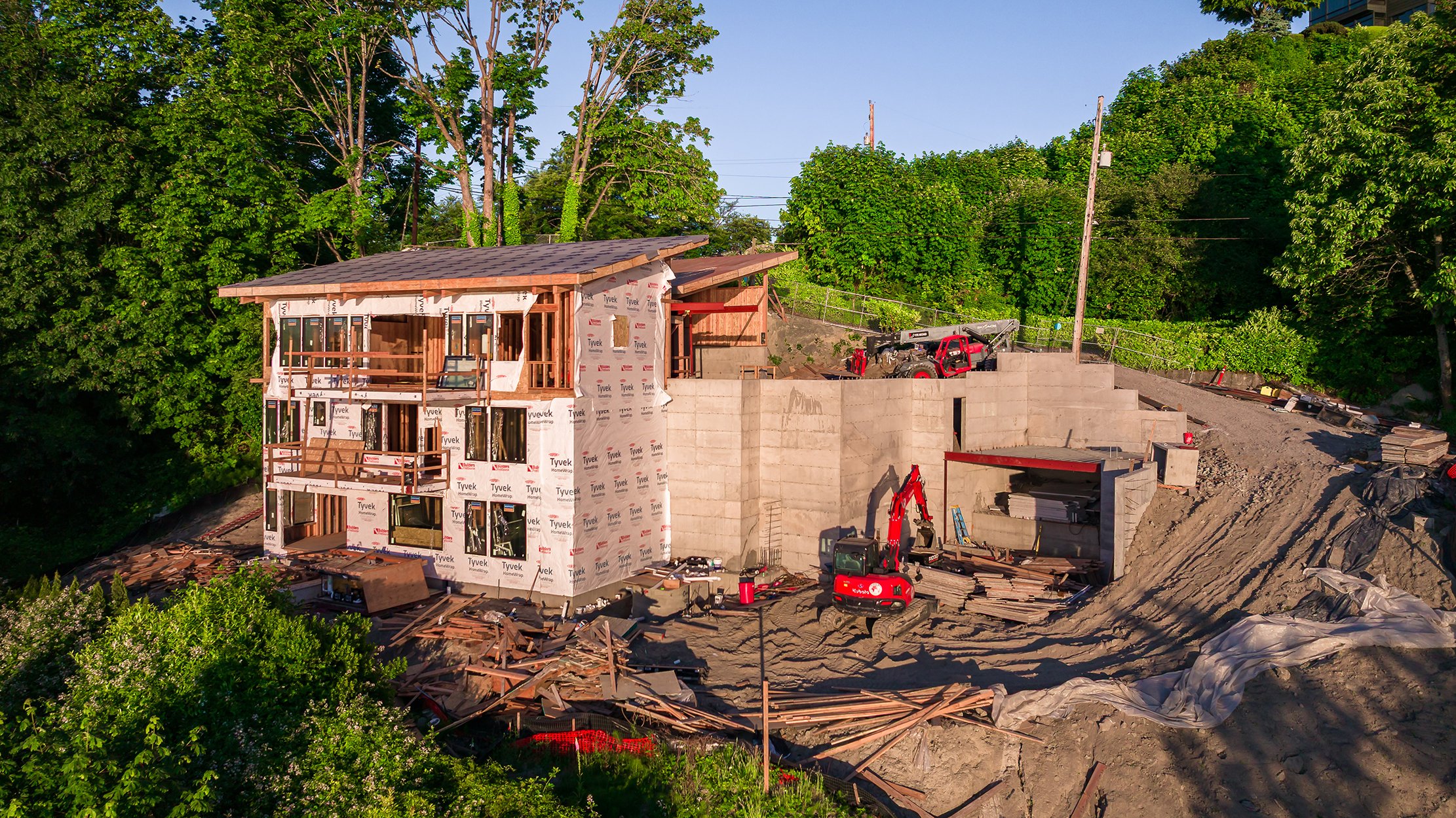 A house under construction on a hillside, with trees in the background, construction equipment, and building materials around.