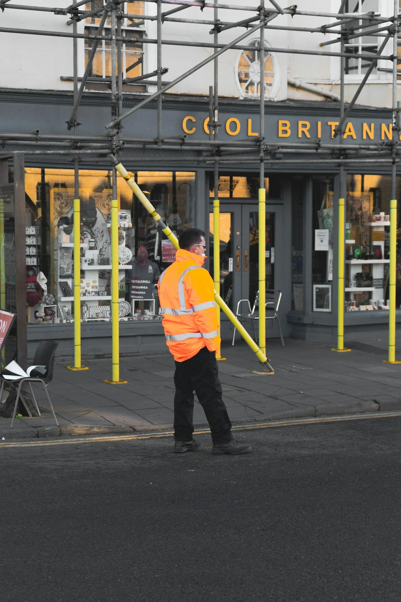 A worker in an orange safety jacket and face mask standing on a city sidewalk in front of a building with scaffolding. The building has a sign reading 'COOL BRITANNIA' and a storefront display with various items inside.