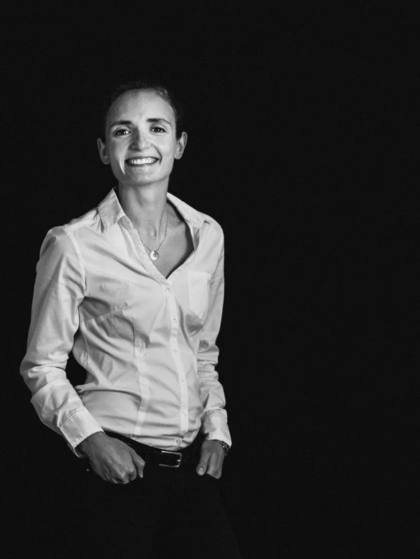 Une femme souriante en chemise blanche, debout contre un fond noir.