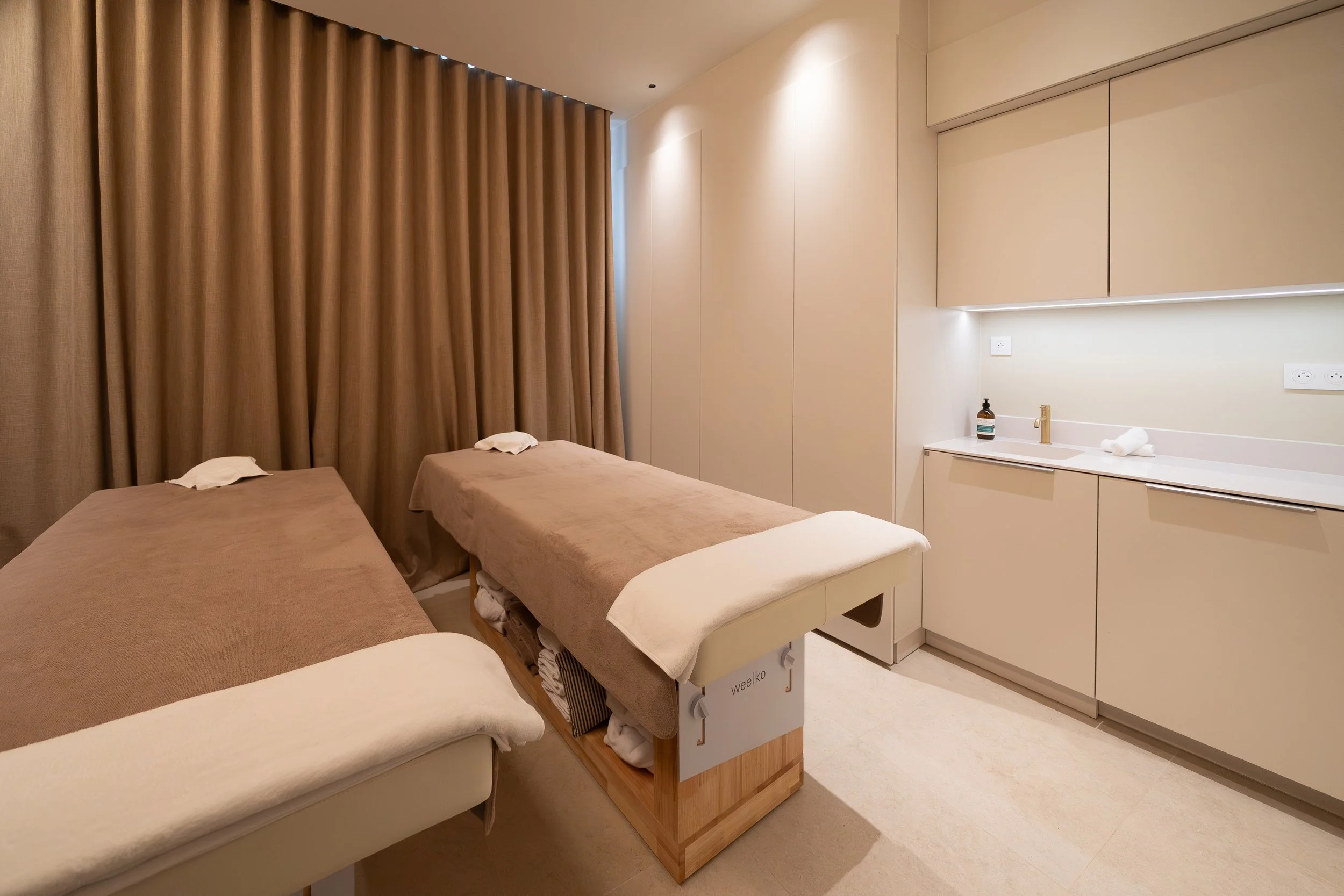 Chambre avec trois lits, rideaux marrons, mobilier blanc, lavage de mains avec savon et serviette. Cabine Duo Spa Beroa.