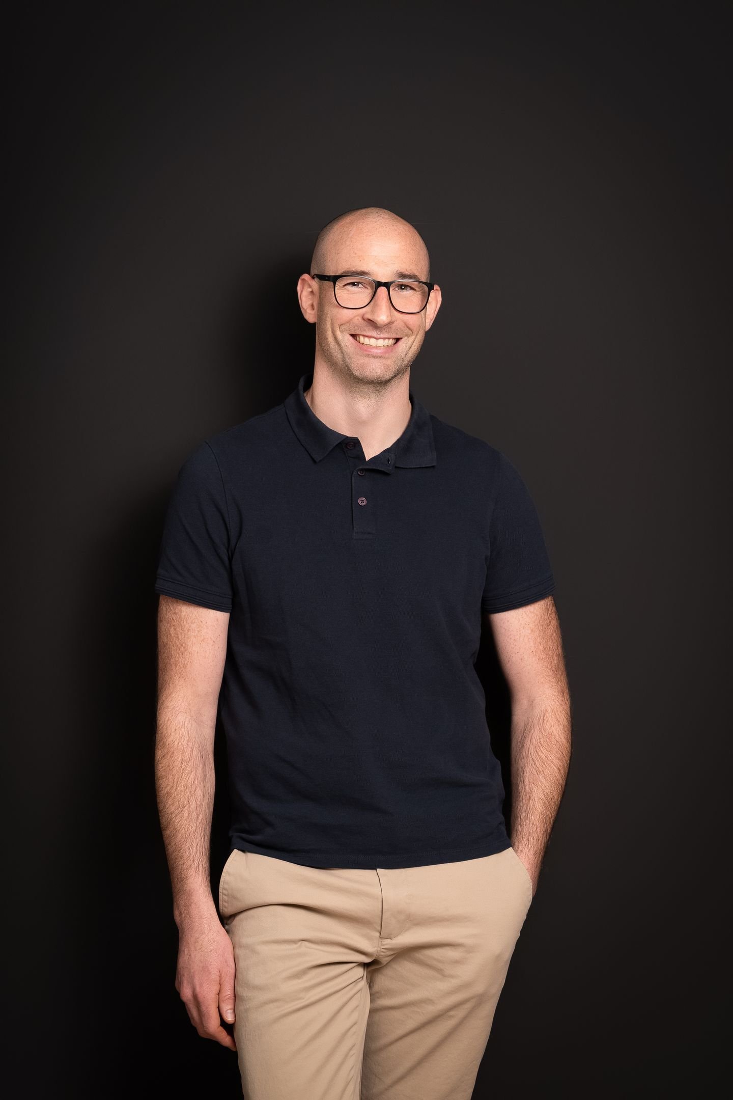 Homme souriant portant des lunettes, vêtu d'un polo noir et de pantalons beige, debout contre un fond noir.