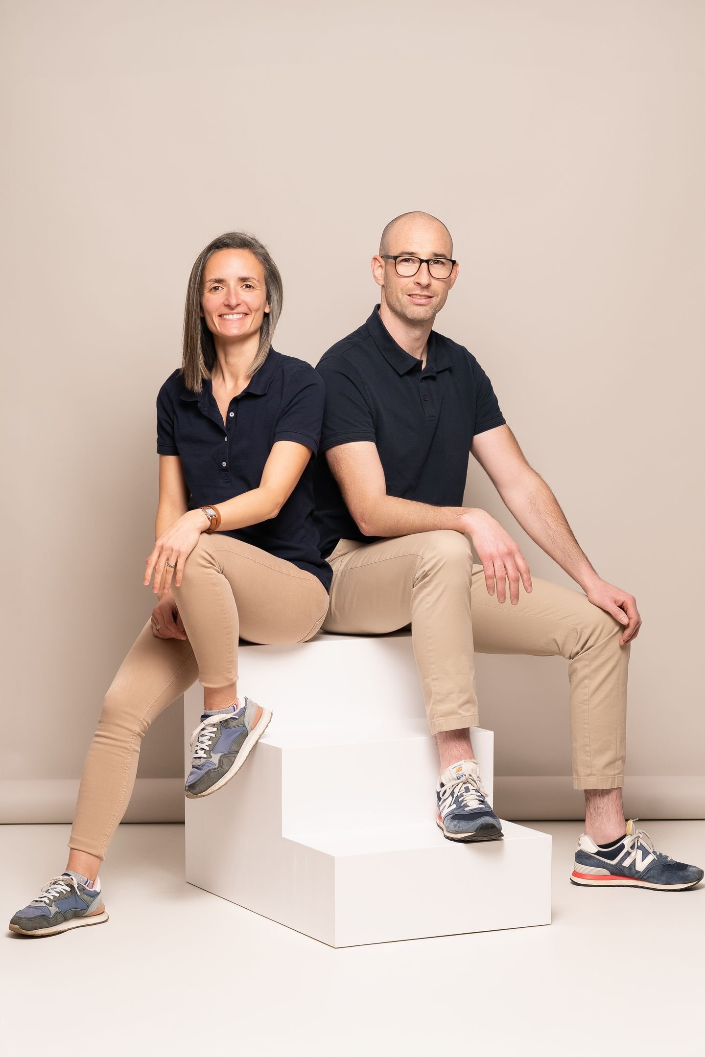Une femme et un homme assis sur une plateforme blanche, tous deux habillés en vêtements décontractés, souriants. Fond beige.