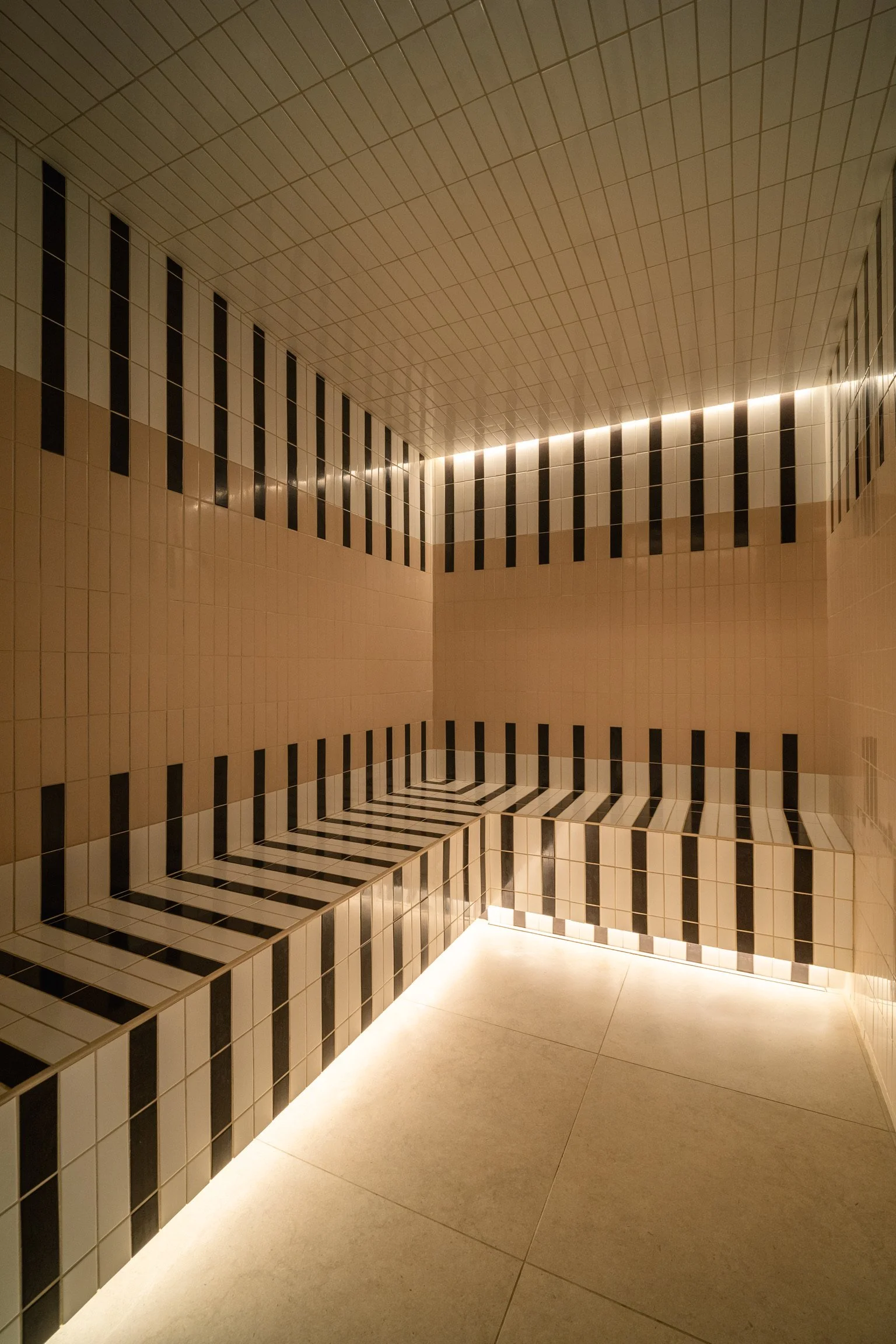 Intérieur d'une pièce avec murs et plafond recouverts de carrelage blanc, noir, clair et beige. Éclairage intégré au plafond créant un effet d'ambiance. Hammam Spa Beroa.