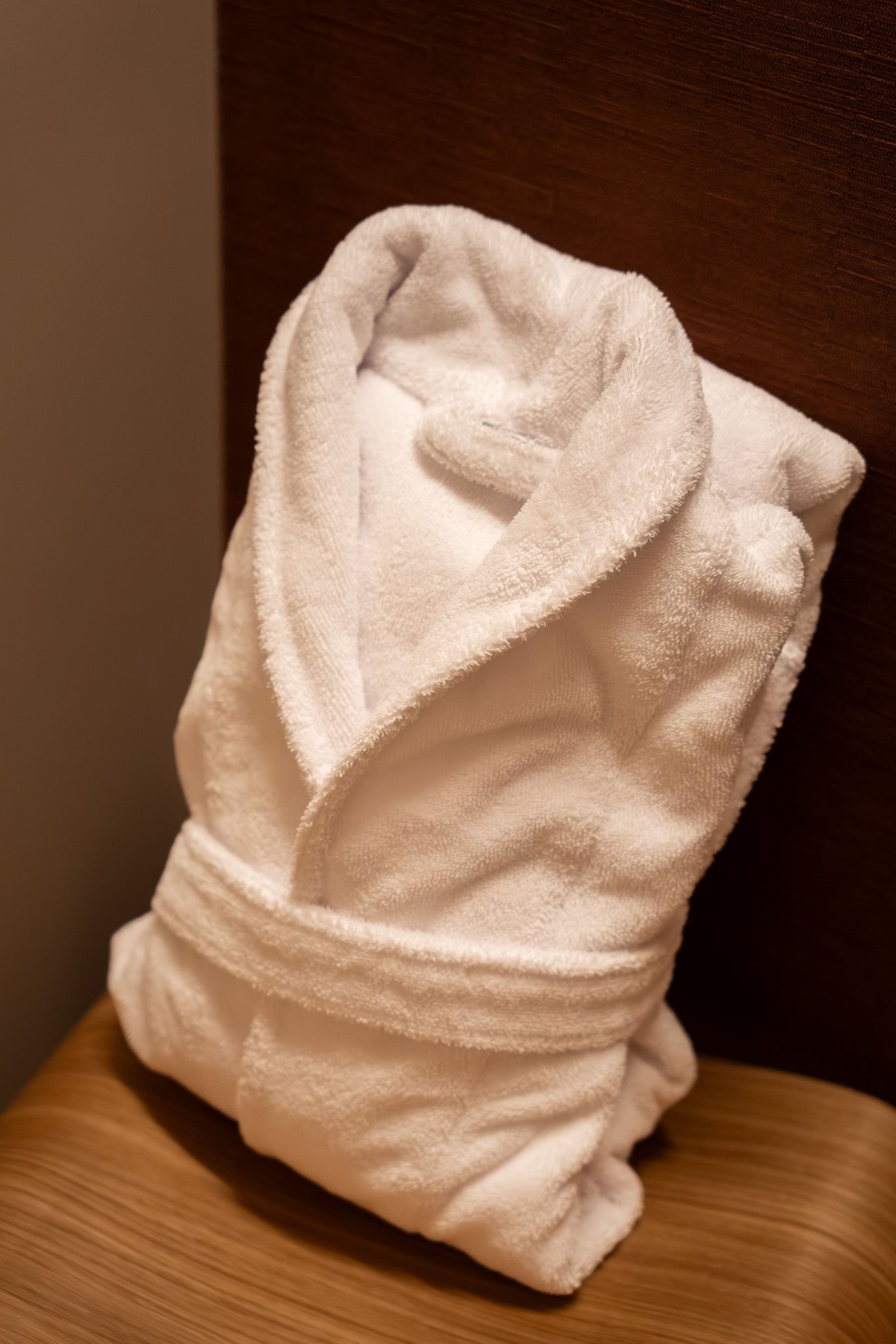 Serviette de bain blanche roulée sur une table en bois, fond d'une tête de lit en bois foncé.