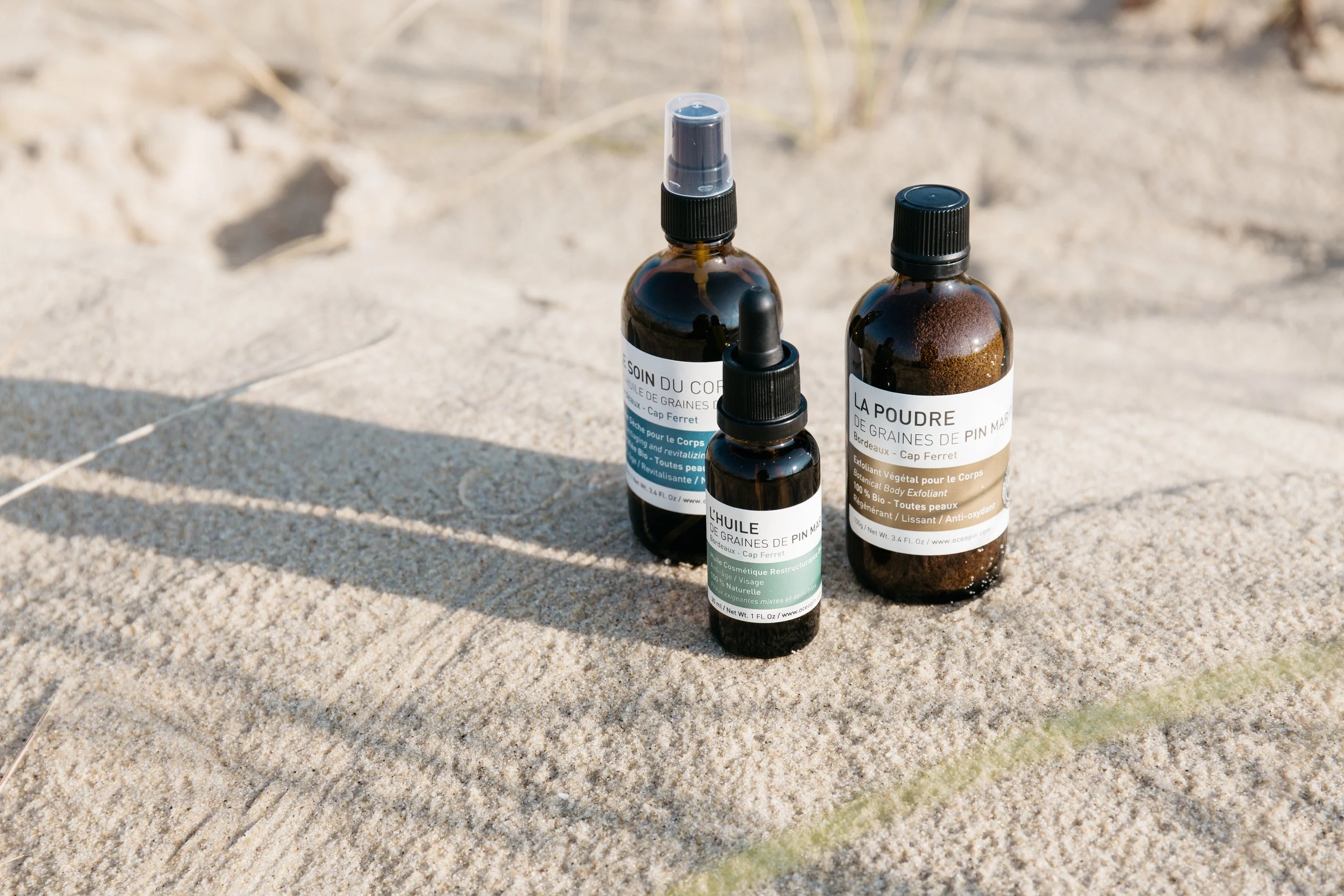 Trois flacons en plastique brun contenant des huiles et exfoliants pour le corps, posés sur du sable en plein air.