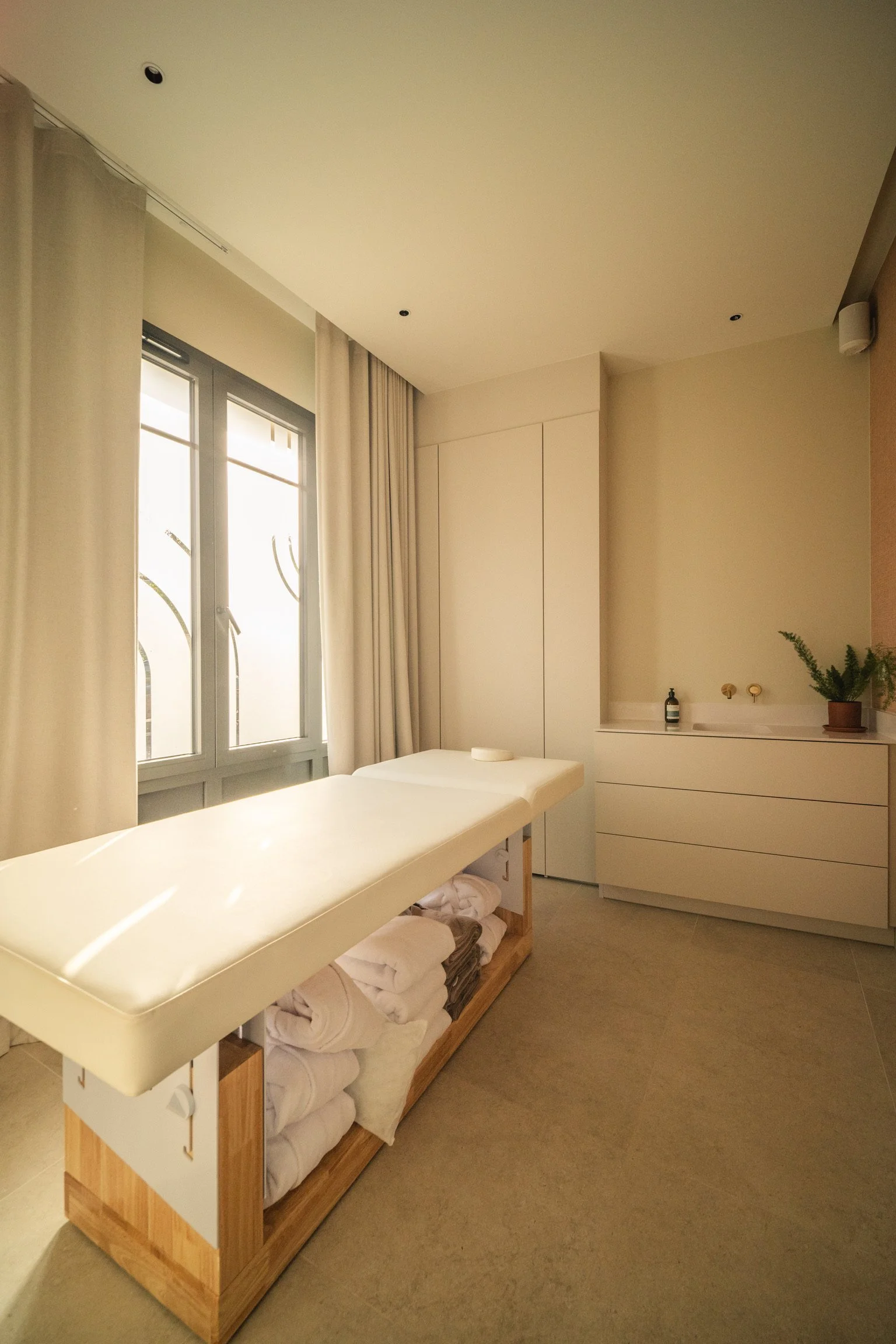 Chambre avec lit, grande fenêtre avec rideaux, meuble blanc, plante dans un pot, bouteille de savon ou lotion sur un meuble. Cabine Solo Spa Beroa.