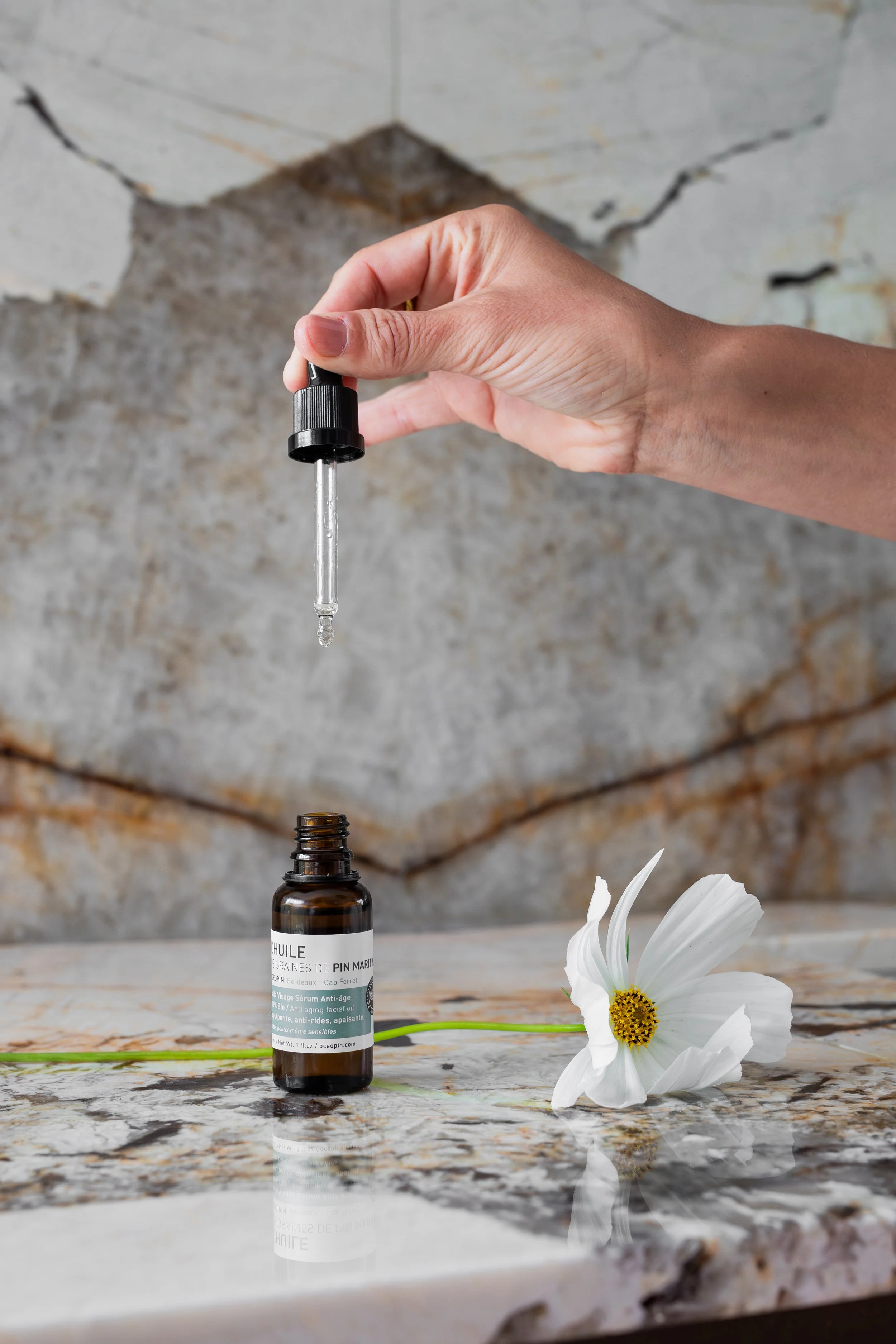 Une main tenant un compte-gouttes avec une goutte d'huile essentielle en dessous, positionnée au-dessus d'un flacon d'huile essentielle de pin maritime. Une fleur blanche repose sur une surface de marbre, avec un fond de mur en pierres.